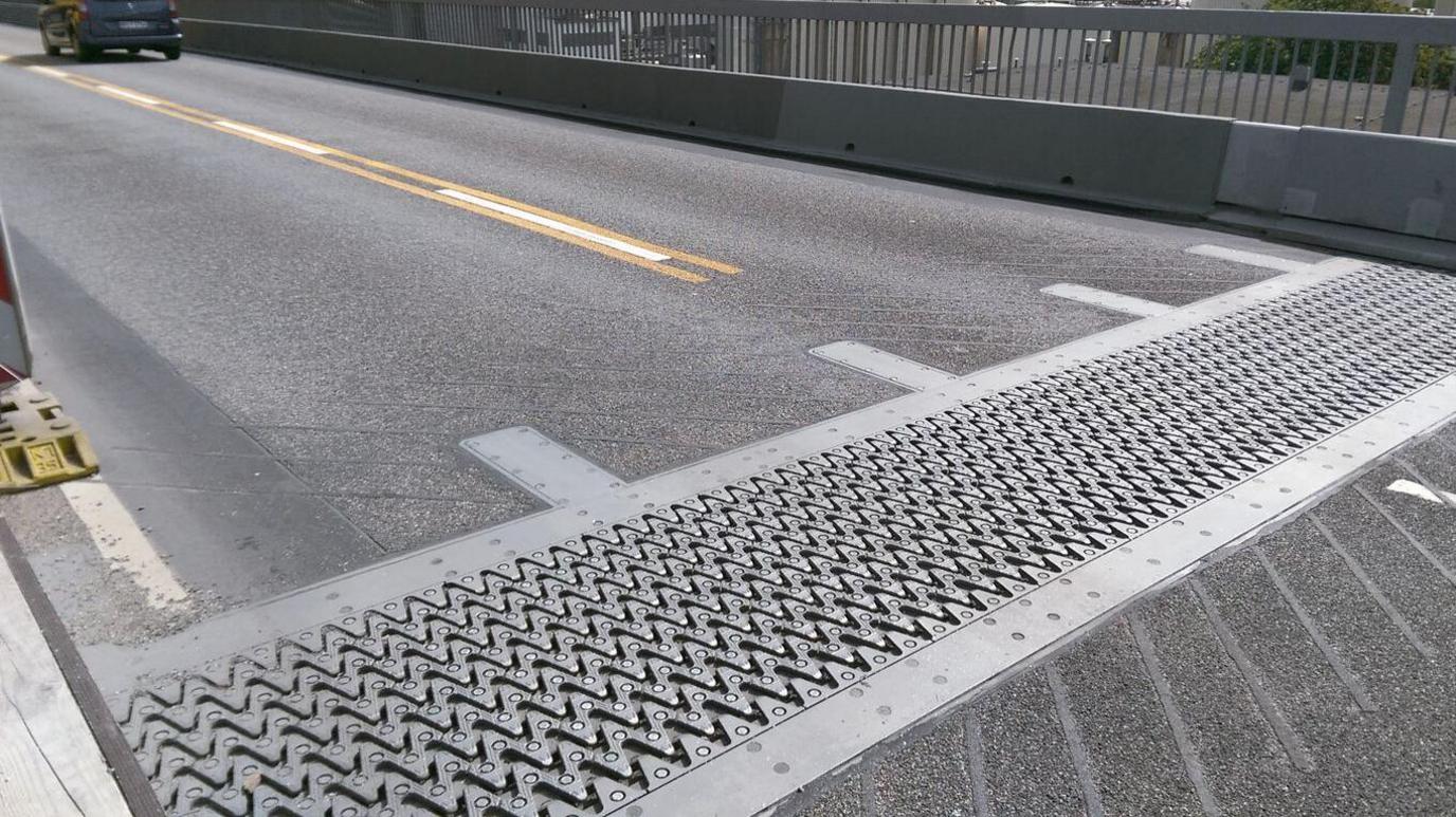 A road from the left side looking down at a silver bridge joint with a zig zag pattern. There are white and yellow painted lines on the bridge and a silver barrier running alongside the road.