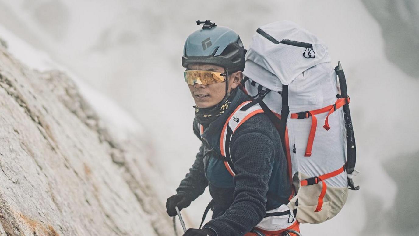 A man in climbing gear wearing a large backpack scales a cliff with a rope