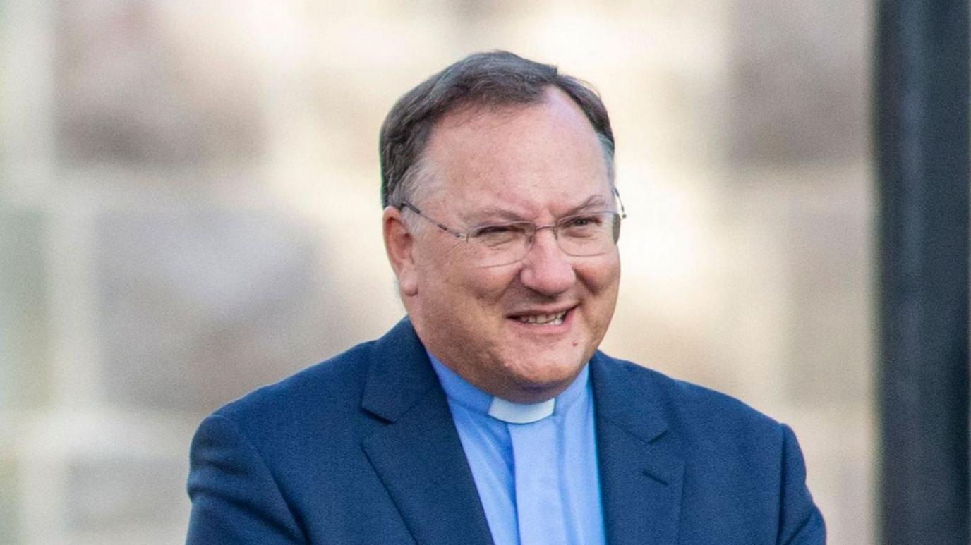 Rev Dr Trevor Gribben, a man with short dark grey hair, wearing a navy suit jacket, a light blue shirt and glasses.