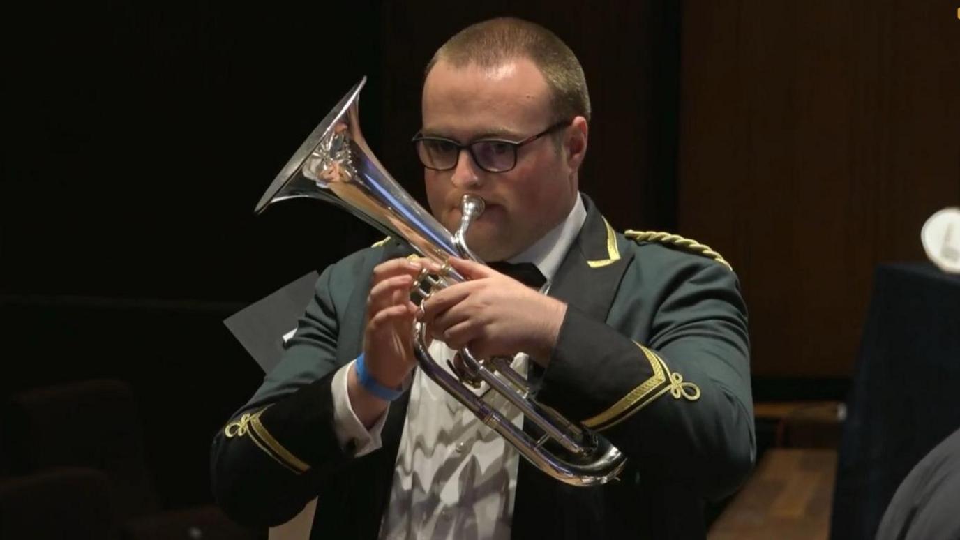 A young man is pictured from the waist up. He is wearing a white shirt and green blazer with gold threaded embellishments. He is holding a silver tenor horn to his mouth.