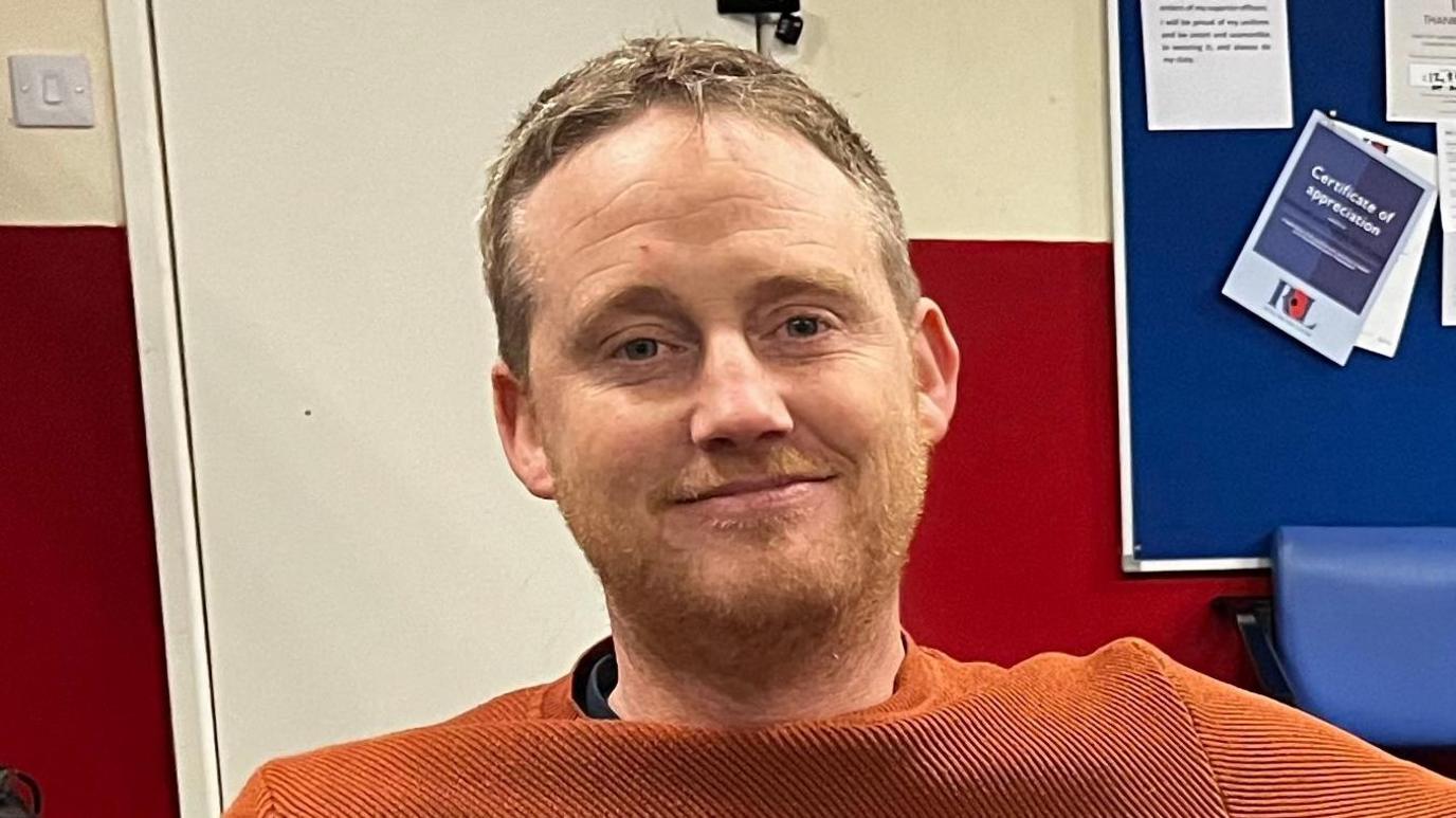 A man sitting in an orange jumper is sitting looking straight to camera. In the background there is a TV on a stand and a noticeboard.