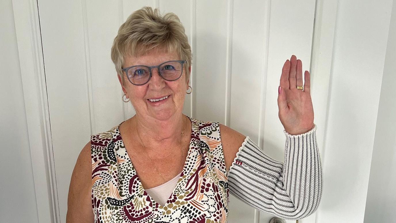 Gilly Windle is stood in front of a white door and is waving at the camera, smiling. She is wearing a wedding ring and has short, light hair and rimmed glasses.