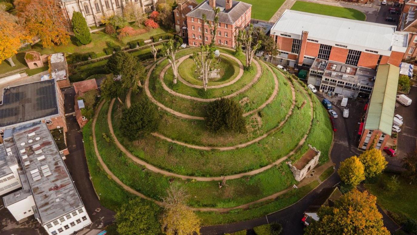 A drone shot of the mound, showing the spiral pathway all the way round to the top. It is mostly covered in grass, but there is the odd tree and a small grotto area at the side. It is surrounded by school buildings, mostly red brick and several storeys high.