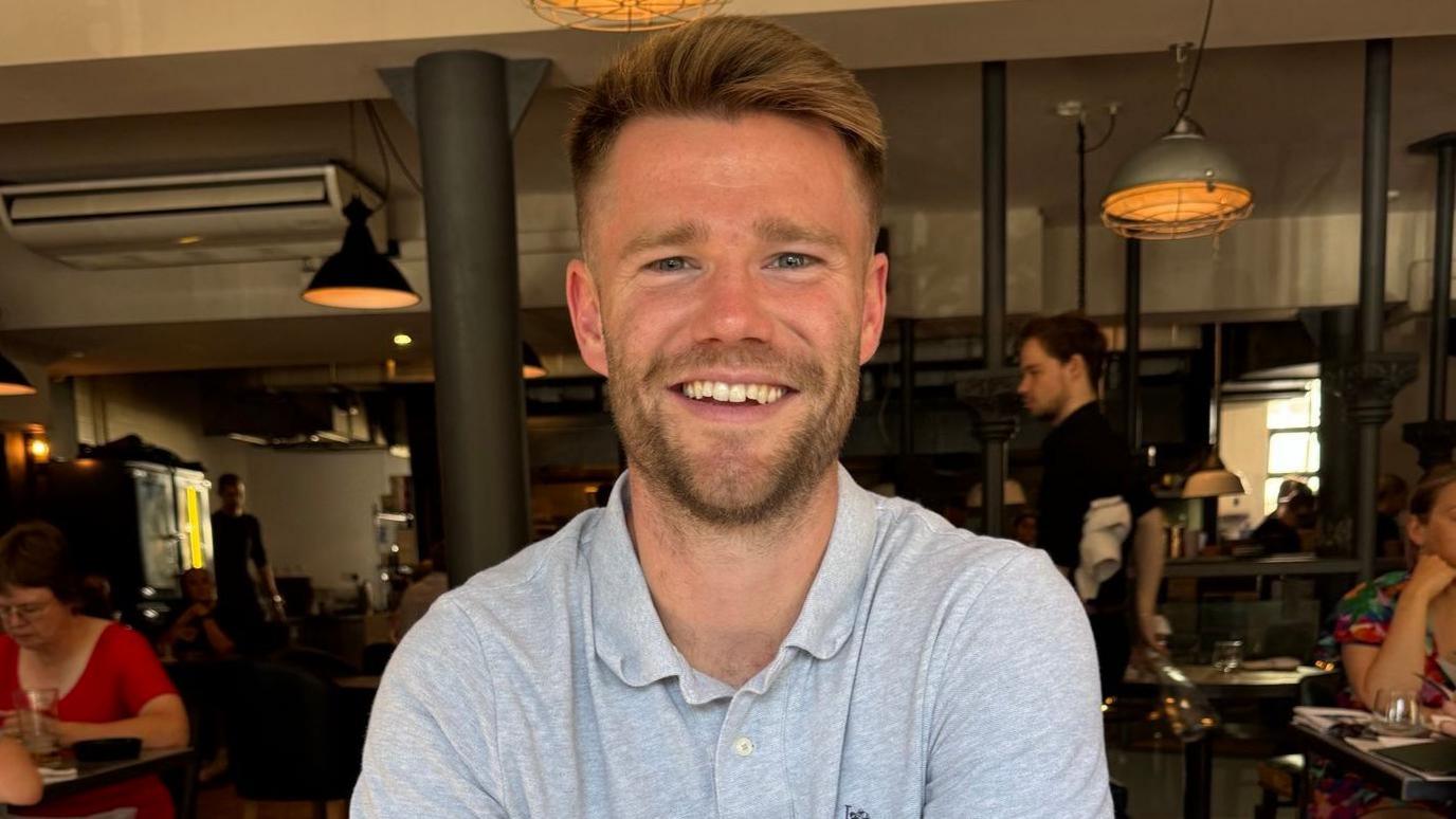 Hamish Douglas, a bearded man in his late 20s or early 30s smiling and wearing a grey polo shirt. He is sat inside a bar or restaurant, with industrial-style lighting behind him. Other customers can be seen at tables nearby.