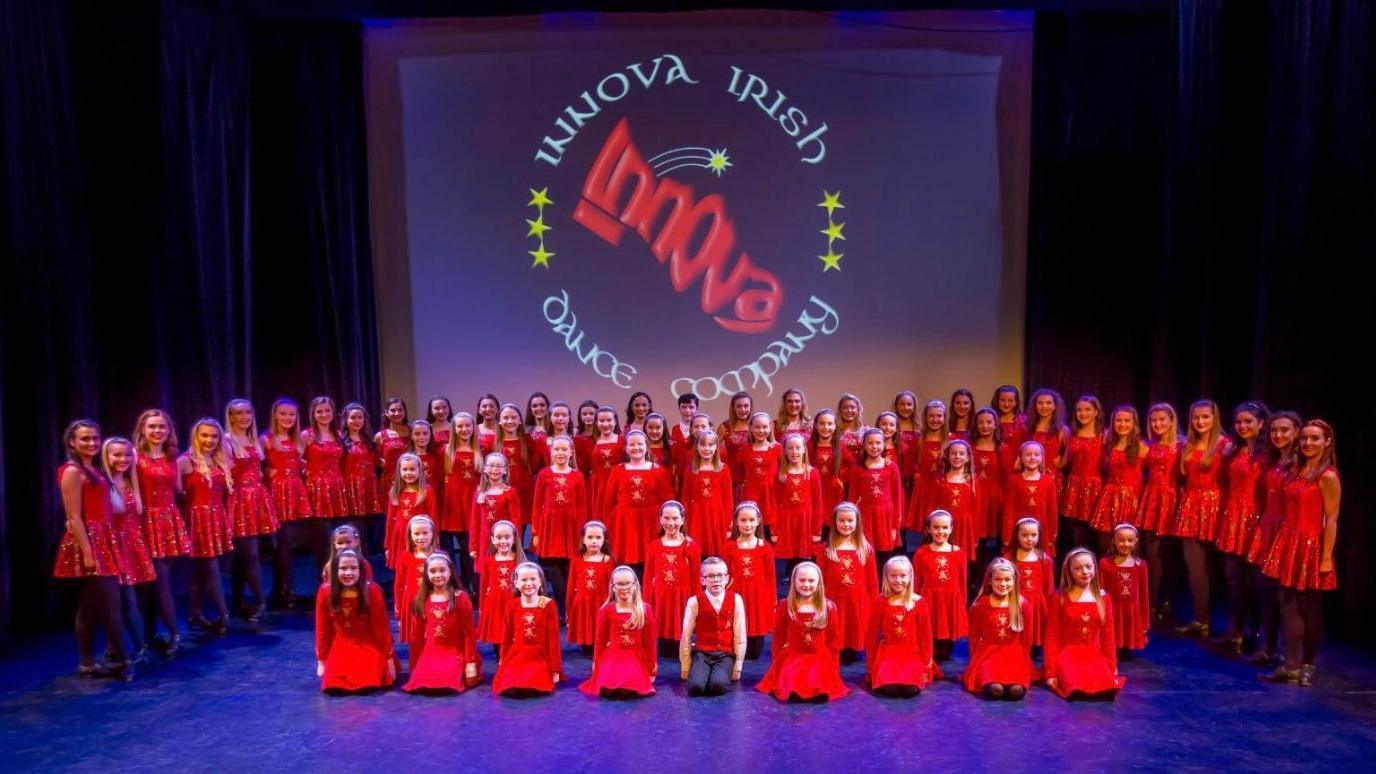 A large group of young dancers from the Innova dance school on stage at the Riverside Theatre. They are all wearing red. The downlighting on stage makes the floor look purple. There is a logo projected on to the back of the curtain saying Innova Irish Dance Company.