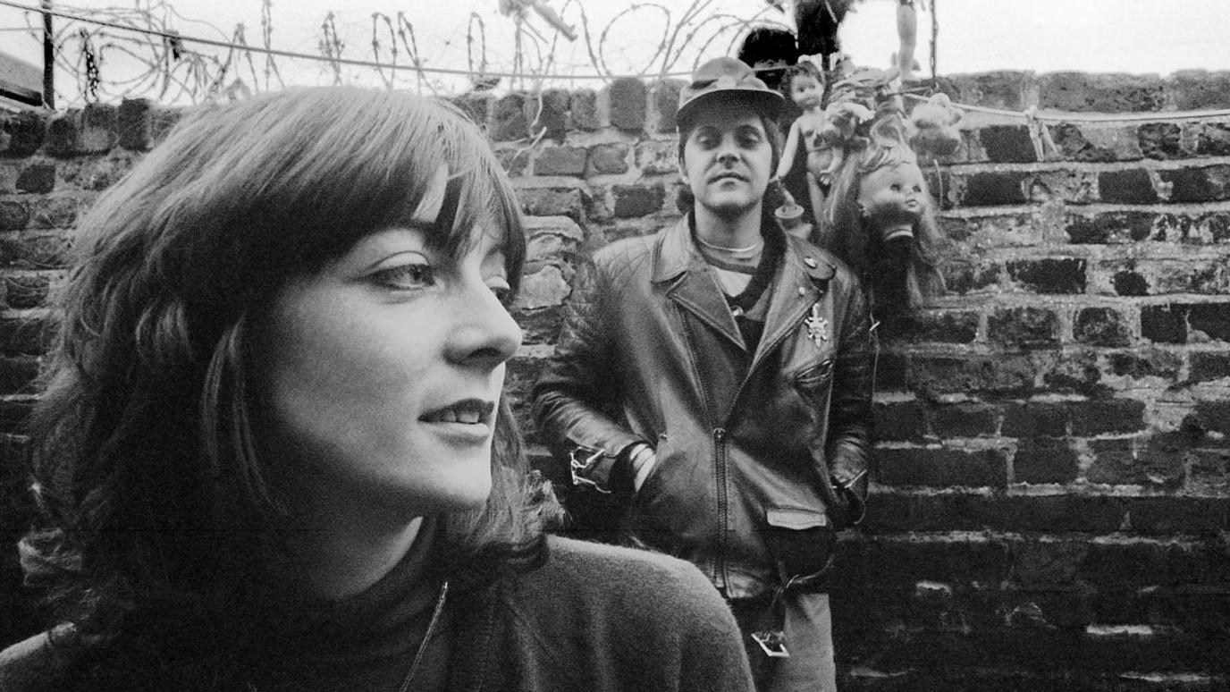 A black and white photo of British performance artist and musician Cosey Fanni Tutti, and British singer-songwriter and musician Genesis P-Orridge of Throbbing Gristle, with a display of dolls' heads in their Hackney garden, protected with barbed wire, in London, England, 1978. Cosey has shoulder length hair and a fringe and is looking to the side and Genesis is wearing a leather jacket and leaning against a brick wall with his hands in his pockets.
