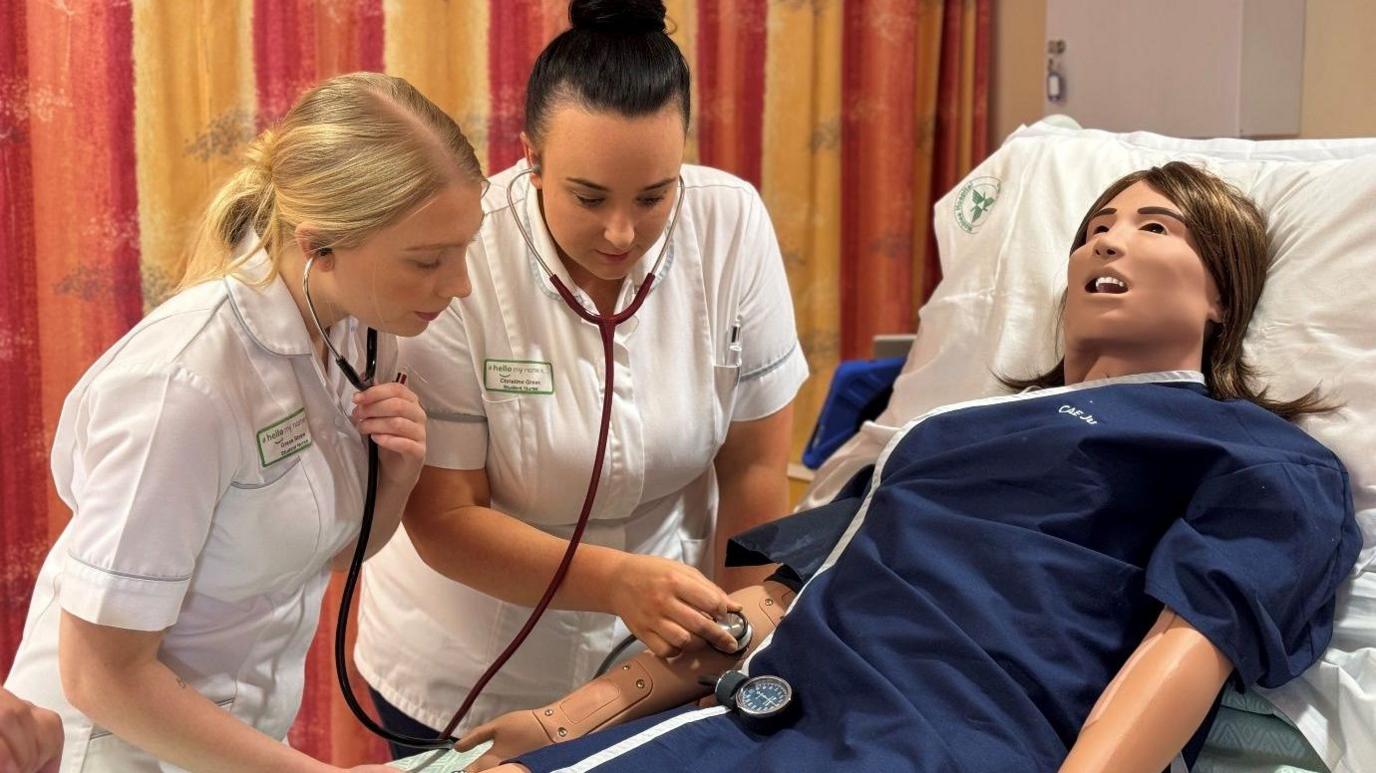 Two young nurses in white uniforms listening to stethoscopes and taking the pulse of a life-like, plastic training mannequin lying in a hospital bed. 