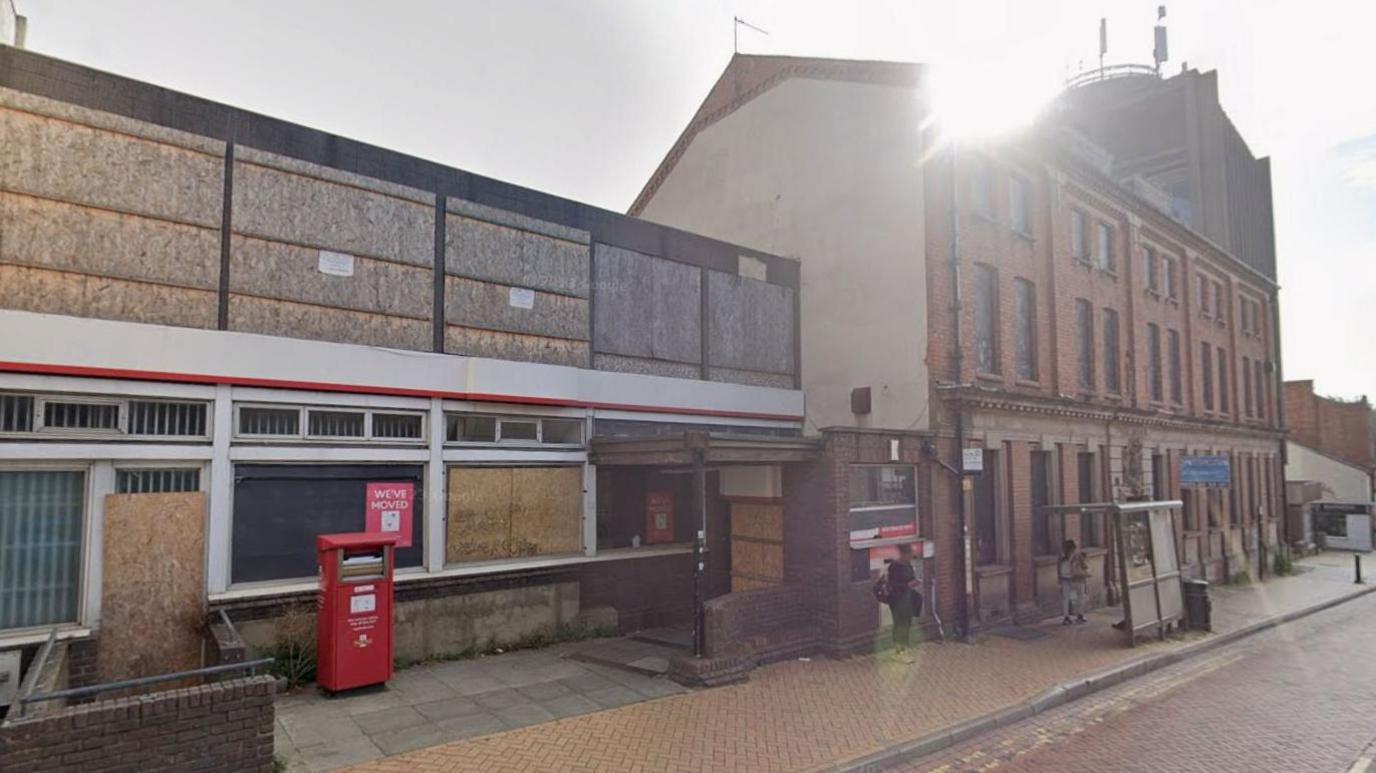 A wider view of the three-storey building which is adjacent to a two-storey more modern concrete building that housed the old post office. All the windows are boarded up with plywood. There is a red post box in front of it and a red poster in one window saying "We've moved".