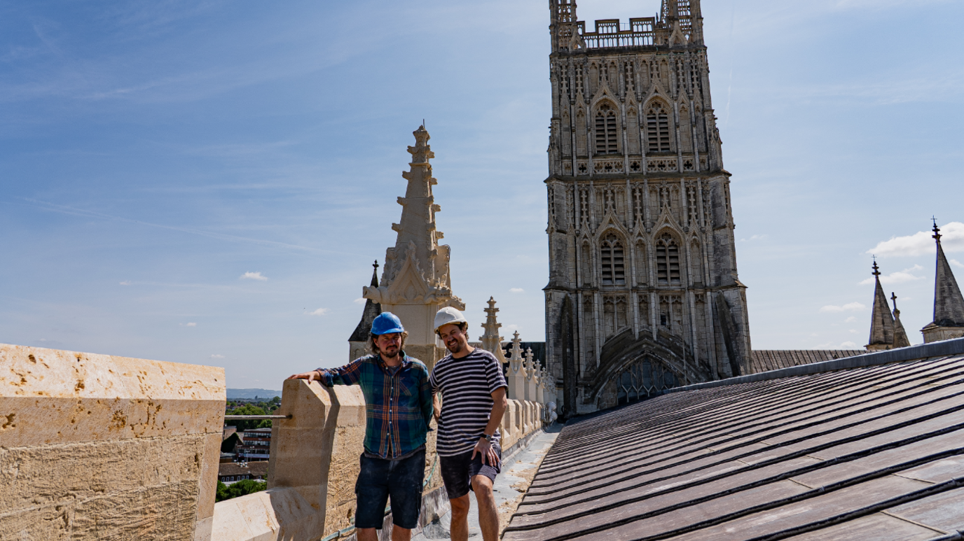 Paul Synan and James Bayliss stand on the roof of Gloucester Cathedral. They are both wearing hard hats and shorts. Mr Synan wears a plaid shirt and Mr Bayliss a stripy top.