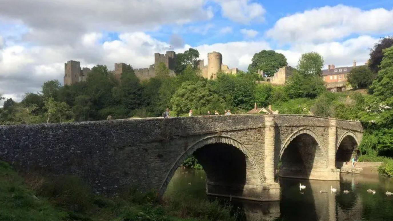An old arched stone bridge over a river with swans below and a castle on the hill in the distance