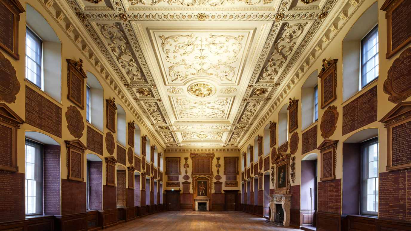 A richly, decorative grand hall, with wooden floors and an intricate gilded ceiling, is surrounded by windows that let in lots of natural light. At the end of the hall is a fireplace, with a painting featured above it.