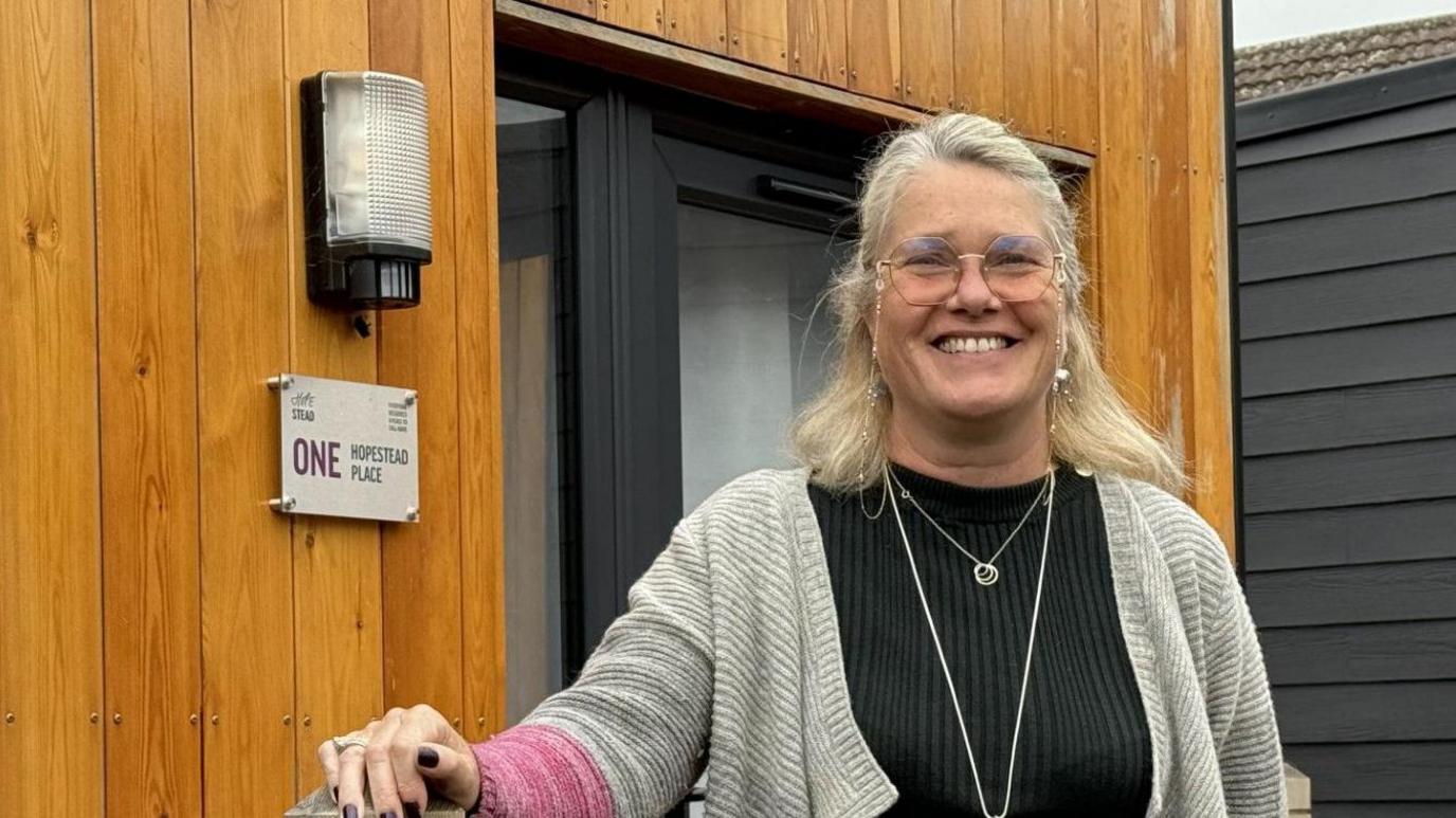 A woman with shoulder-length blondy hair wearing glasses, a grey cardigan over a black jumper. She is smiling into the camera and standing in front of a wooden 'pod' for homeless. 