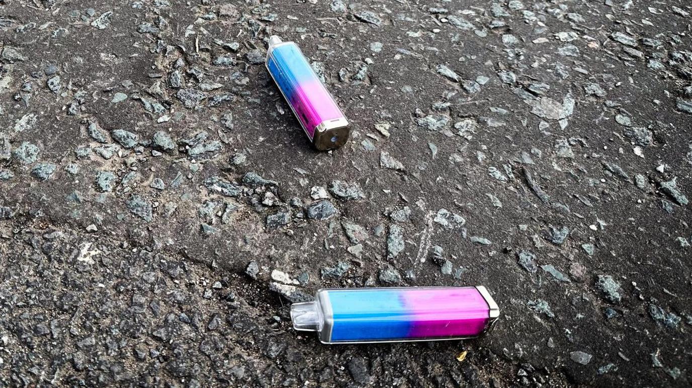 Two single-use vapes on a concrete path or road. They are plastic on the outside, and blue and pink in colour.