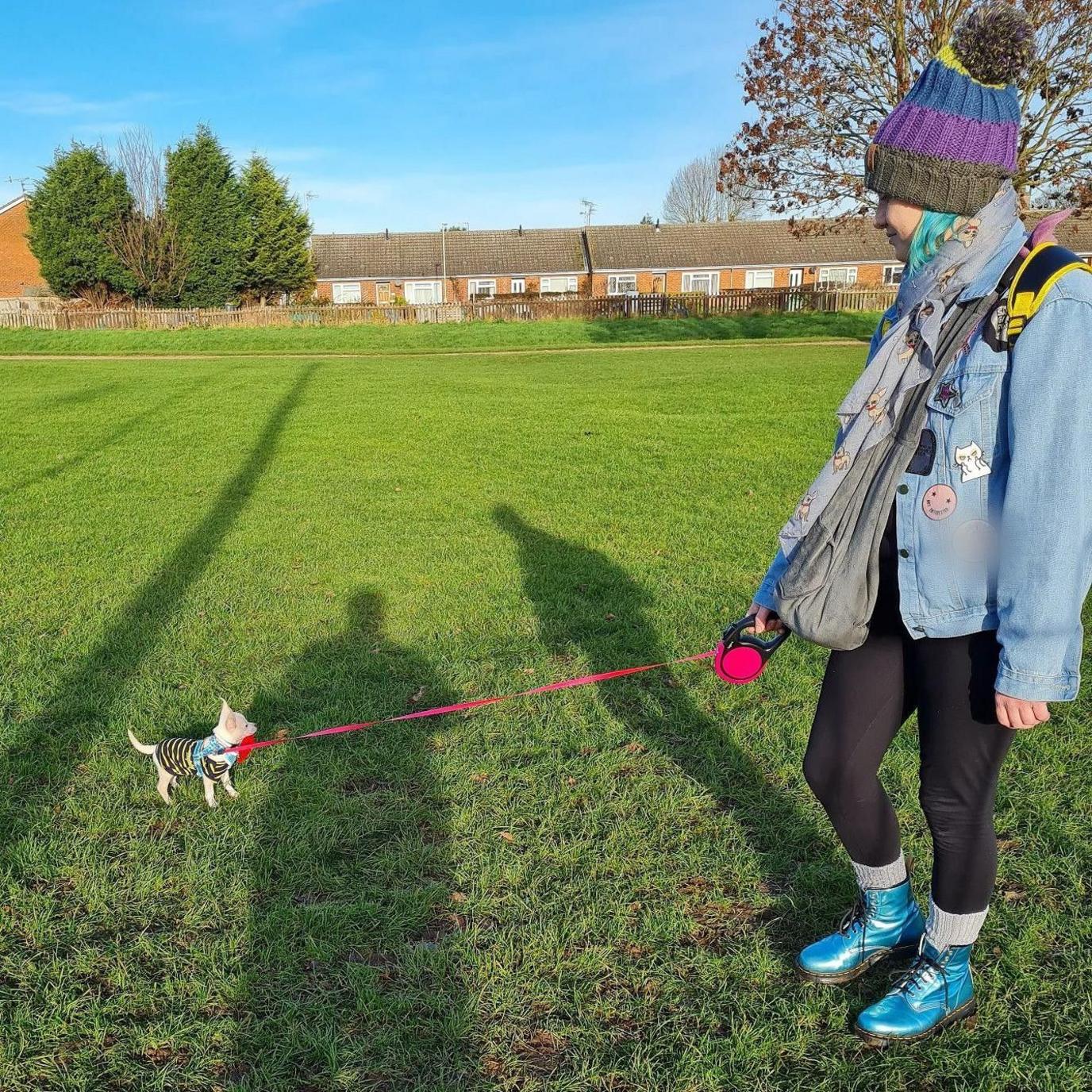 A small dog on a brightly coloured lead being held by a woman in a bobble hat, scarf and jacket, and wearing some snazzy teal Doc Martens