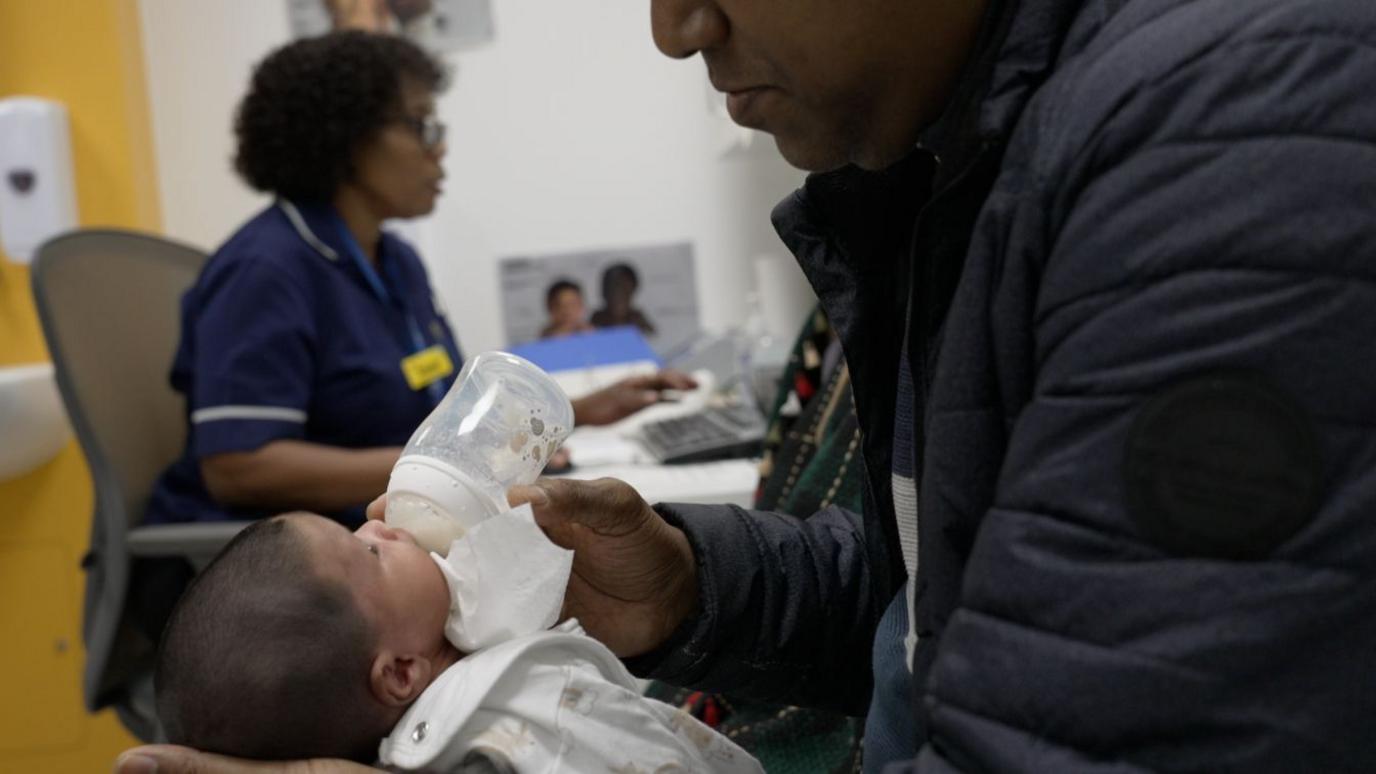 A man is bottle-feeding a baby while Irene works in the background. She is seated at a desk in a clinical setting.