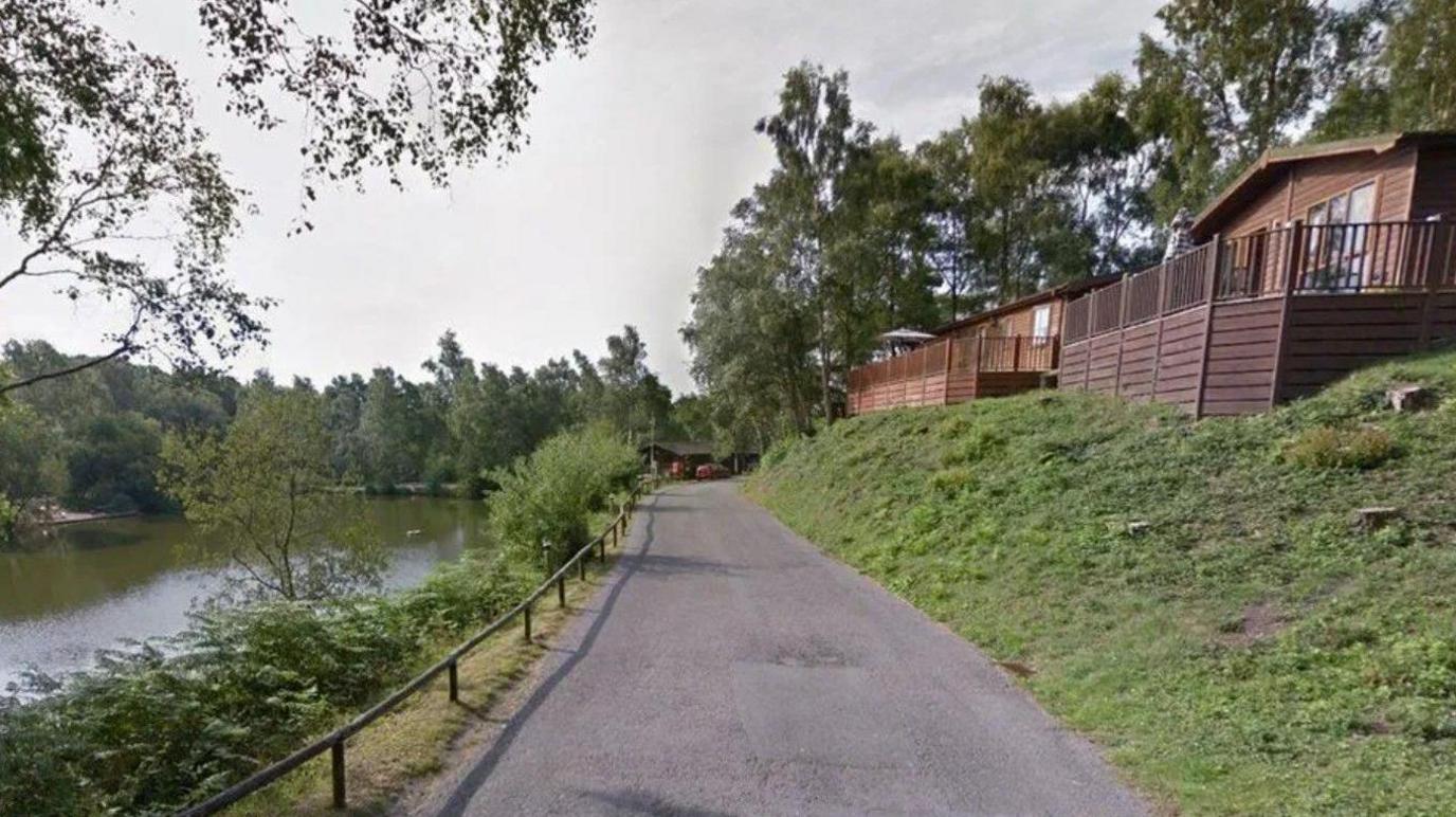 A narrow tarmac road running between a lake which is down a slope to the left and a row of timber cabins up on a bank to the right