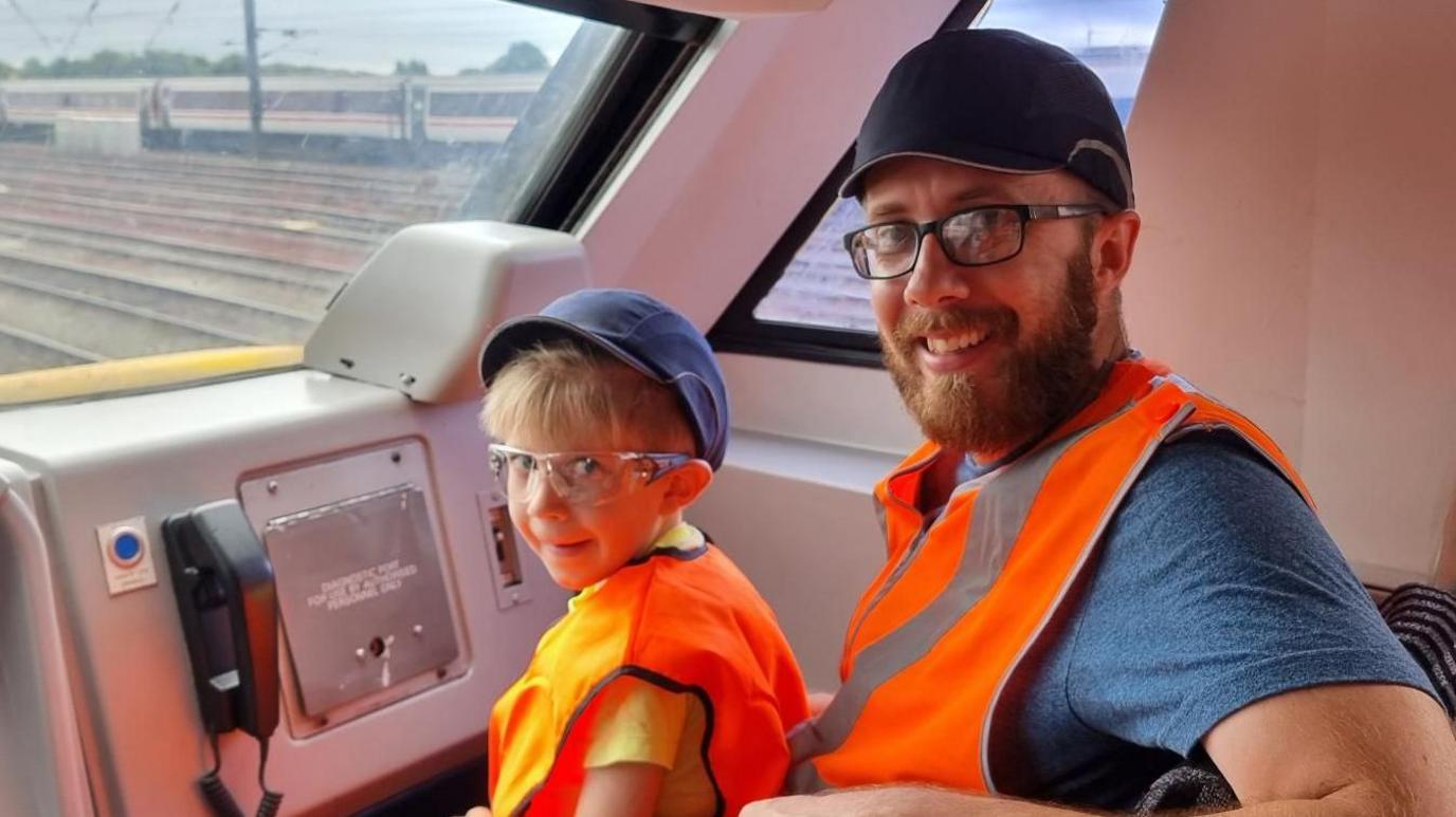 Ashley in a blue top and orange fluorescent hi-vis jacket and blue cap. He is wearing dark rimmed glasses. Sitting with him is Austin. He is also wearing an orange fluorescent hi-vis jacket with glasses on and a blue cap. 