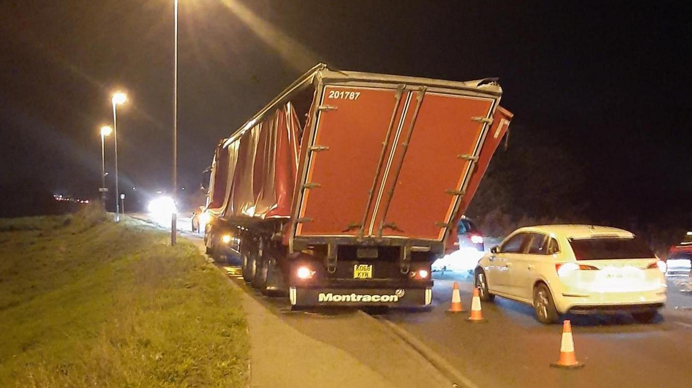 A damaged lorry at the side of the road. It is red and parts of its trailer are damaged and hanging off. The lorry has been coned off. Cars are passing on one side. The shot has been taken in the dark and headlights and streetlights are shining.