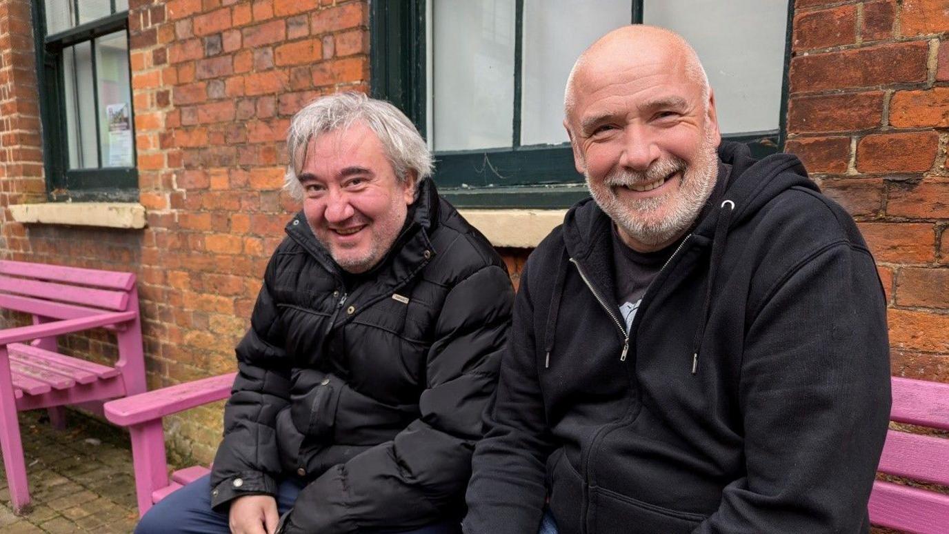 Two men, Lee and Steve, are sitting on a bright pink bench smiling at the camera. Lee has grey stubble and grey hair, he is wearing a black jacket and blue trousers. Steve has a shaved head and white and grey cropped beard. He is wearing a black hoody over a black T-shirt. 