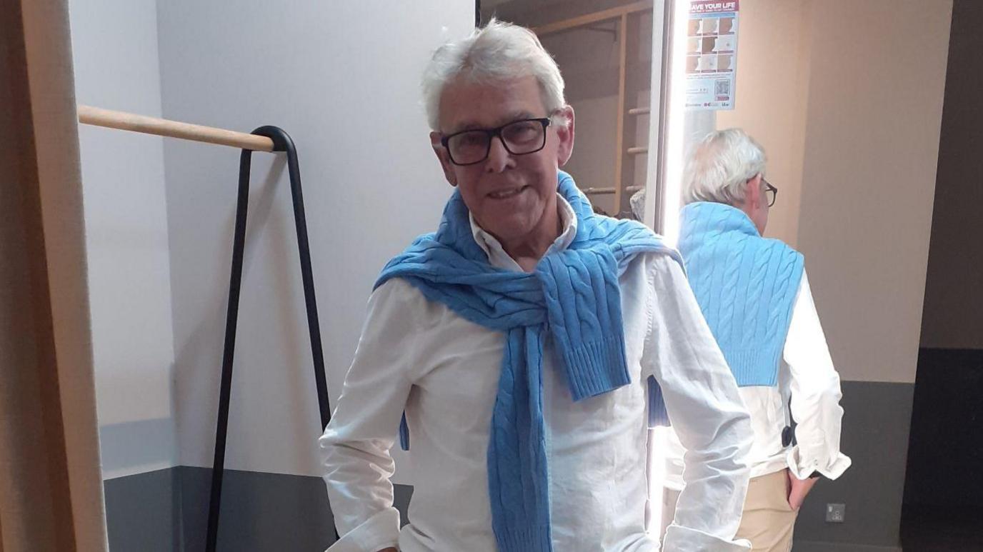 Roger is posing with his hands in his tanned trousers pockets wearing a white shirt and blue cable knit around his neck. 