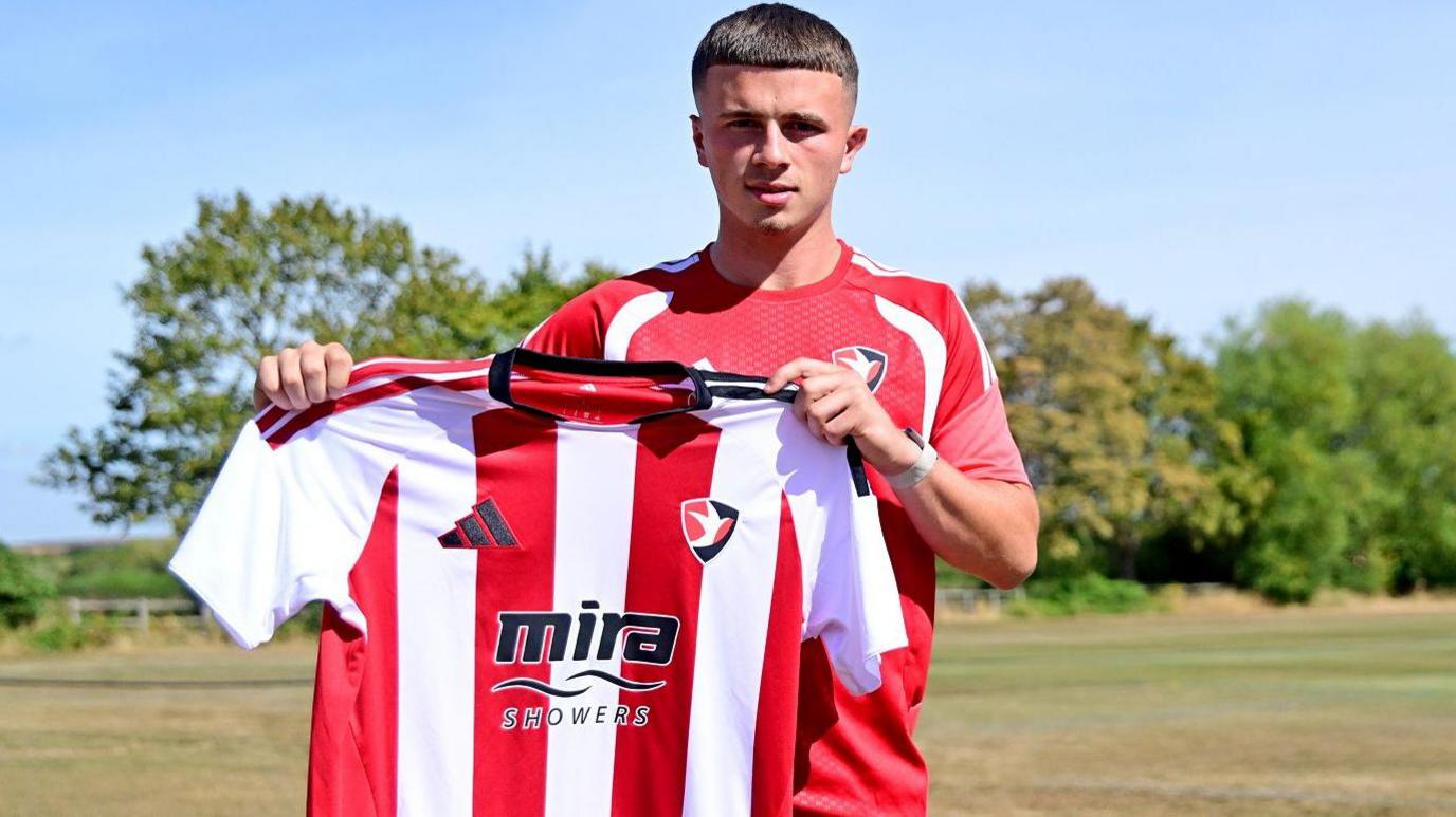 Jake Evans in red Cheltenham Town training top holds up a red and white striped Cheltenham kit with grass and trees behind him.