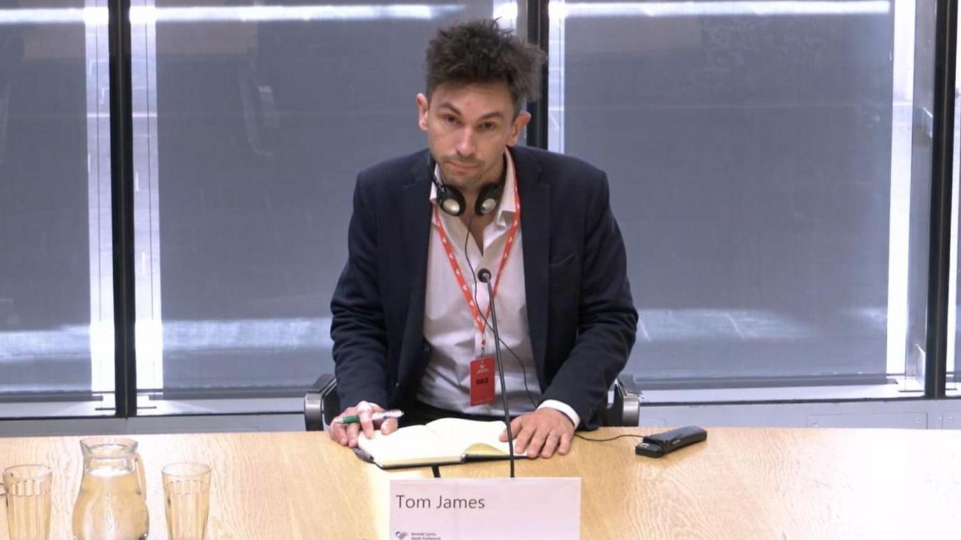 Thomas James, appearing at a Senedd committee meeting, wearing an open necked shirt and a suit jacket, with a laynard and a pair of translation headphones