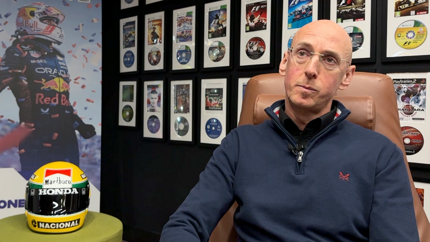 Ian is sat in an office with gaming images and discs on the wall behind him. He is next to a F1 helmet. He is wearing a blue top and glasses.