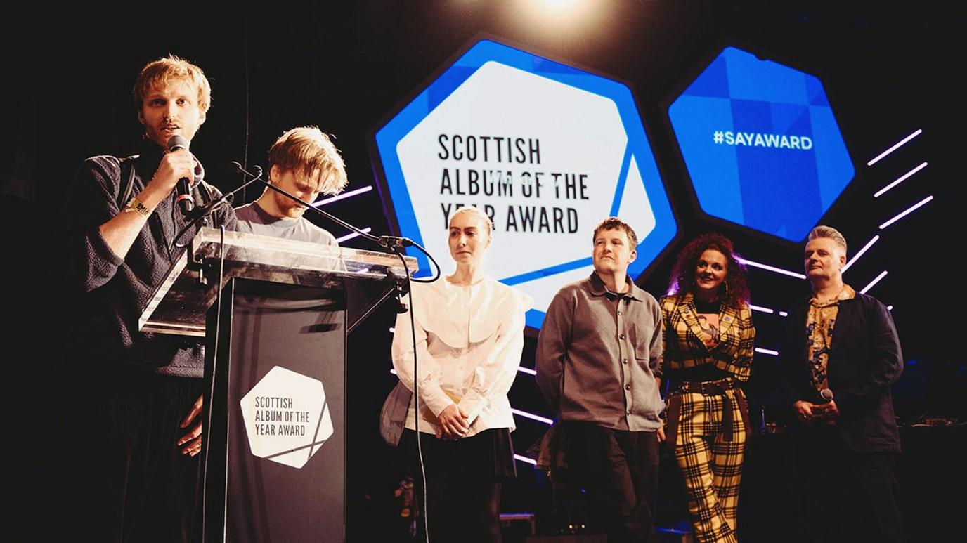 Three members of redolent, two male and one female, stand at a podium in front of a large awards logo with other guests at the side of the stage