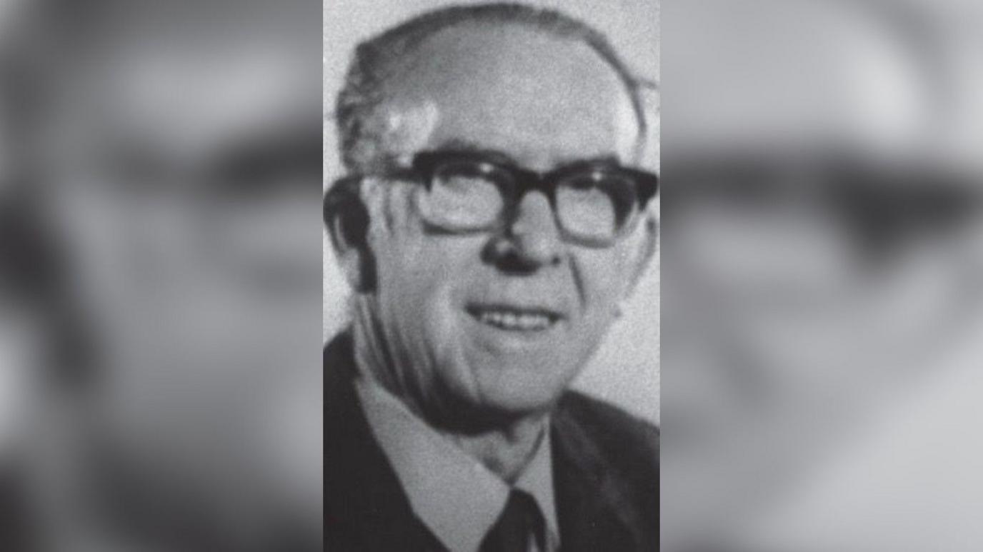 A black and white image of a man in late life with short hair and large black glasses