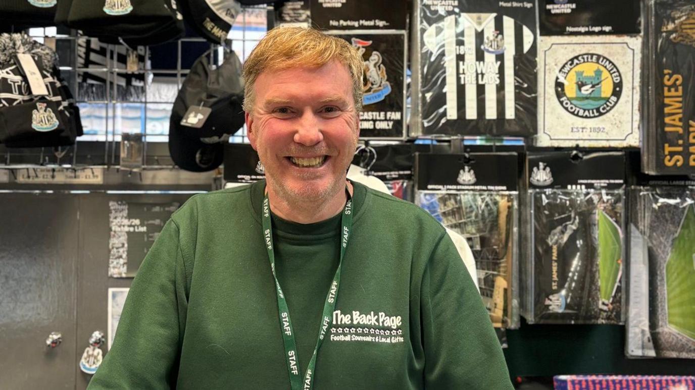 Gary Oliver smiling into the camera. He has light, short hair swept to the side. He has light stubble on his face and is wearing a green jumper with The Back Page logo on the chest. He is standing in front of a wall of Newcastle memorabilia.