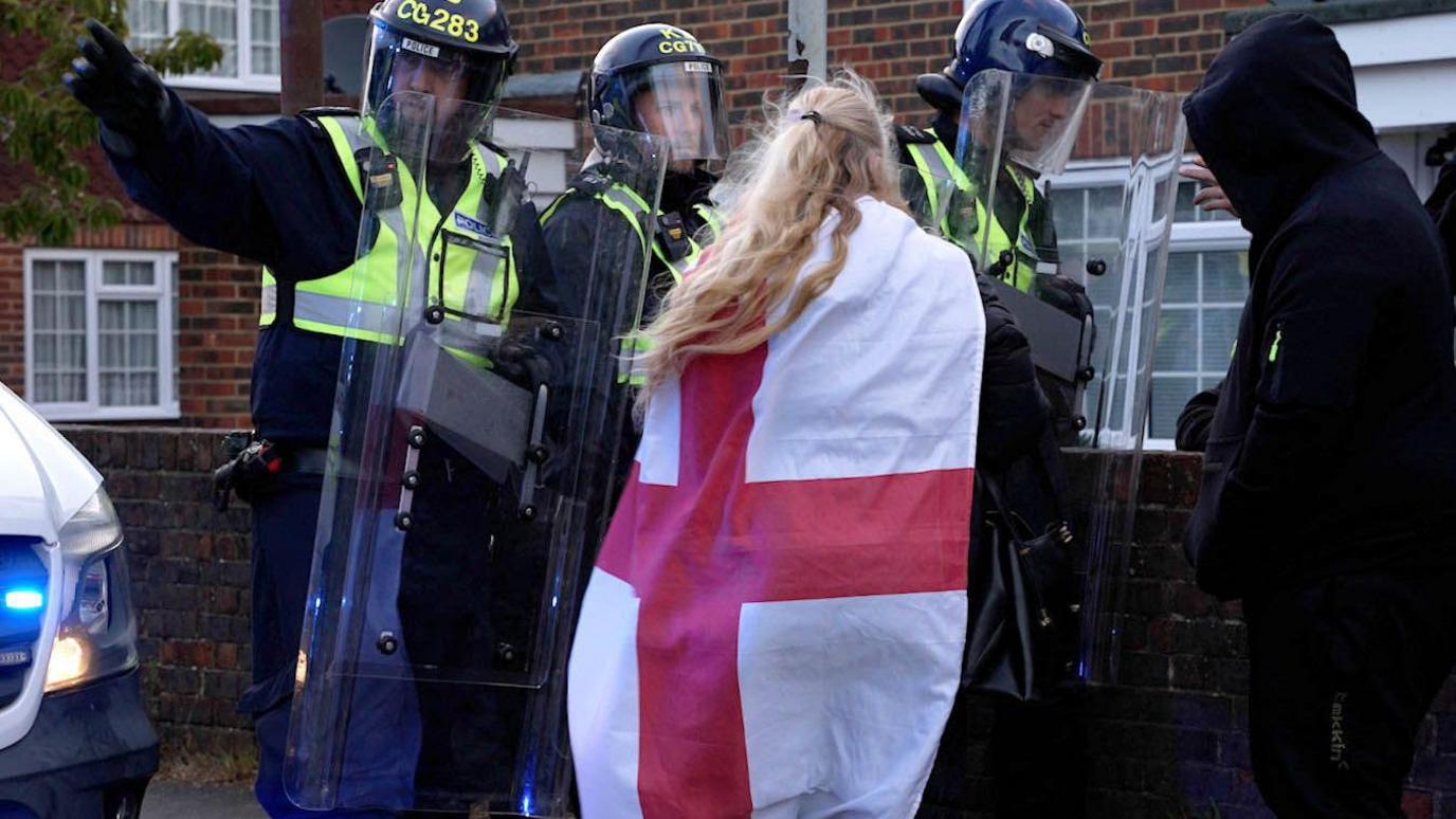 Police officers wearing helmets and holding clear riot shields stand on a residential street, facing members of the public near brick houses and a parked police vehicle.