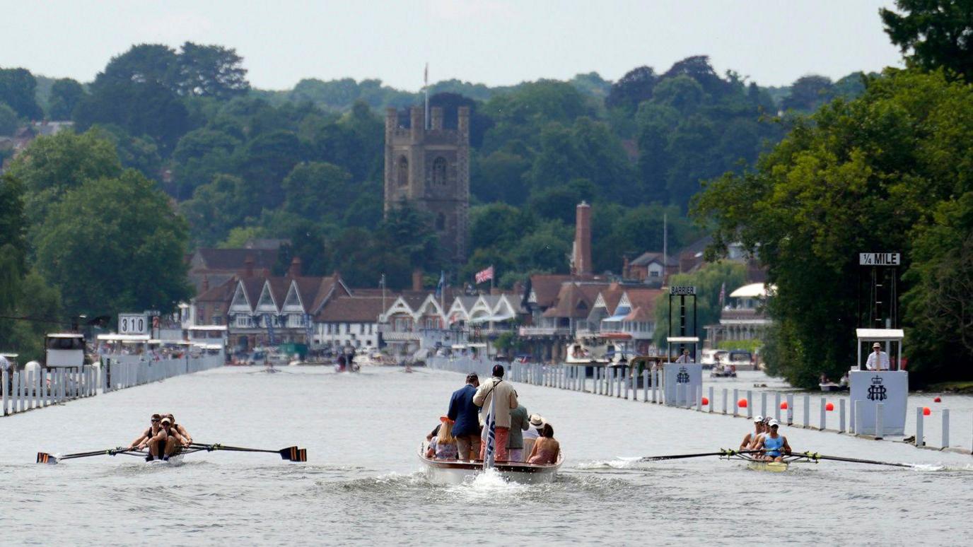 Two rowing boats and a motor boat moving down a river with a row of boat houses, a church and trees in the background.