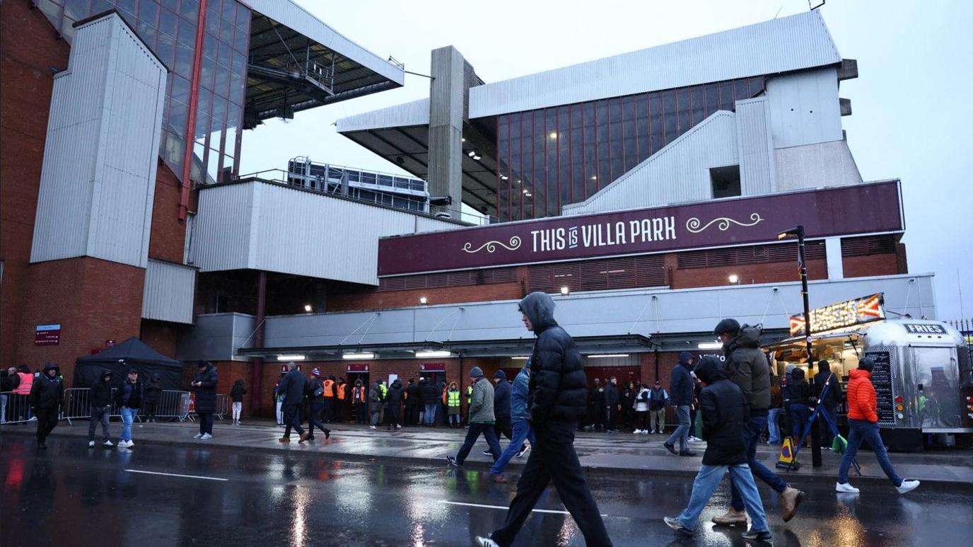 General view of fans outside Aston Villa's Villa Park stadium. It is raining as people ahead with the stadium and a food outlet in the background. There is a sign that reads: "This is Villa Park".