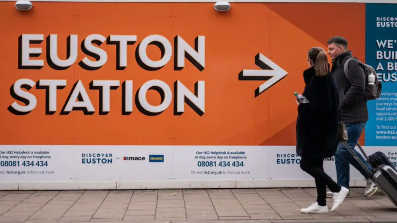 Passengers with suitcases walk past a billboard