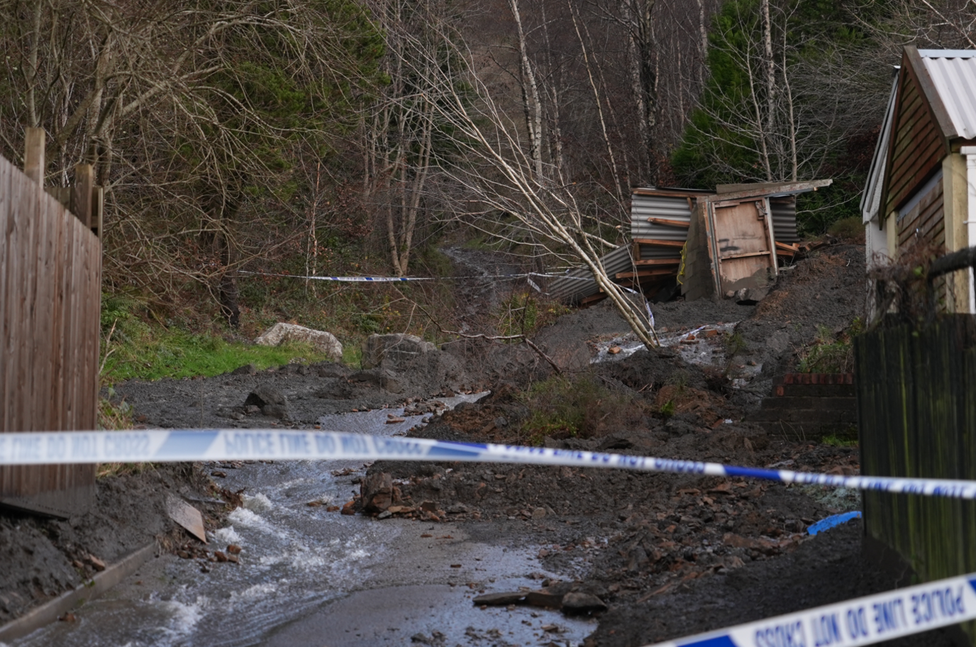 Police tap cordons off a street, there are mounds of rubble and mud. There is debris from small sheds as well as water coming down the road