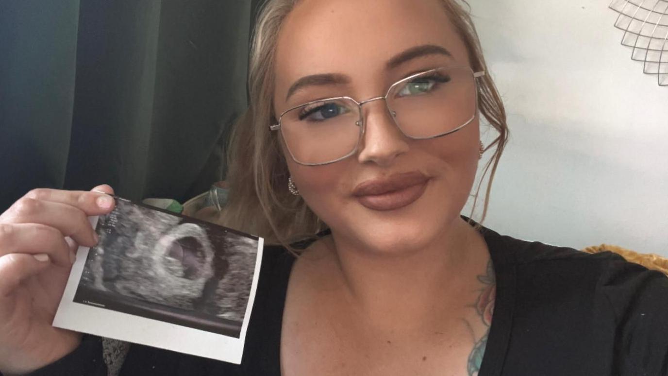 Natasha is smiling at the camera, wearing a black top and glasses, with long blonde hair, and holding a print-out of her pregnancy scan