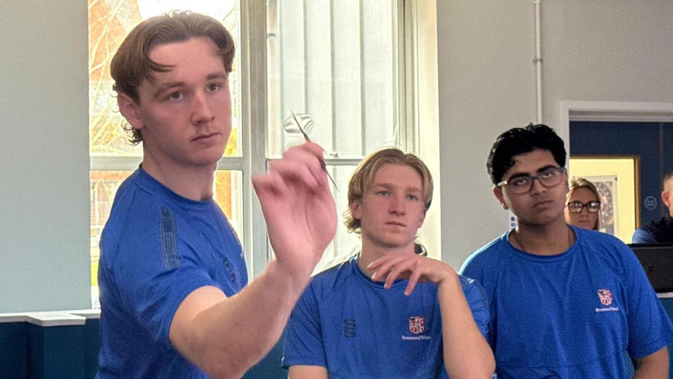 Three boys in blue tops staring past the camera, one of them has a dart poised as if he is about to throw it