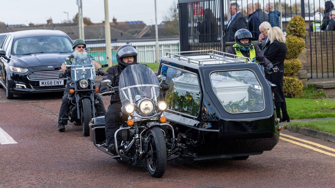 Mr Dingwall's coffin being carried in a black hearse sidecar attached to a motorbike. His brother, Robert is riding the motorcycle. Another biker, with a large white beard is riding behind. People are standing at the side of the road to pay their respects.