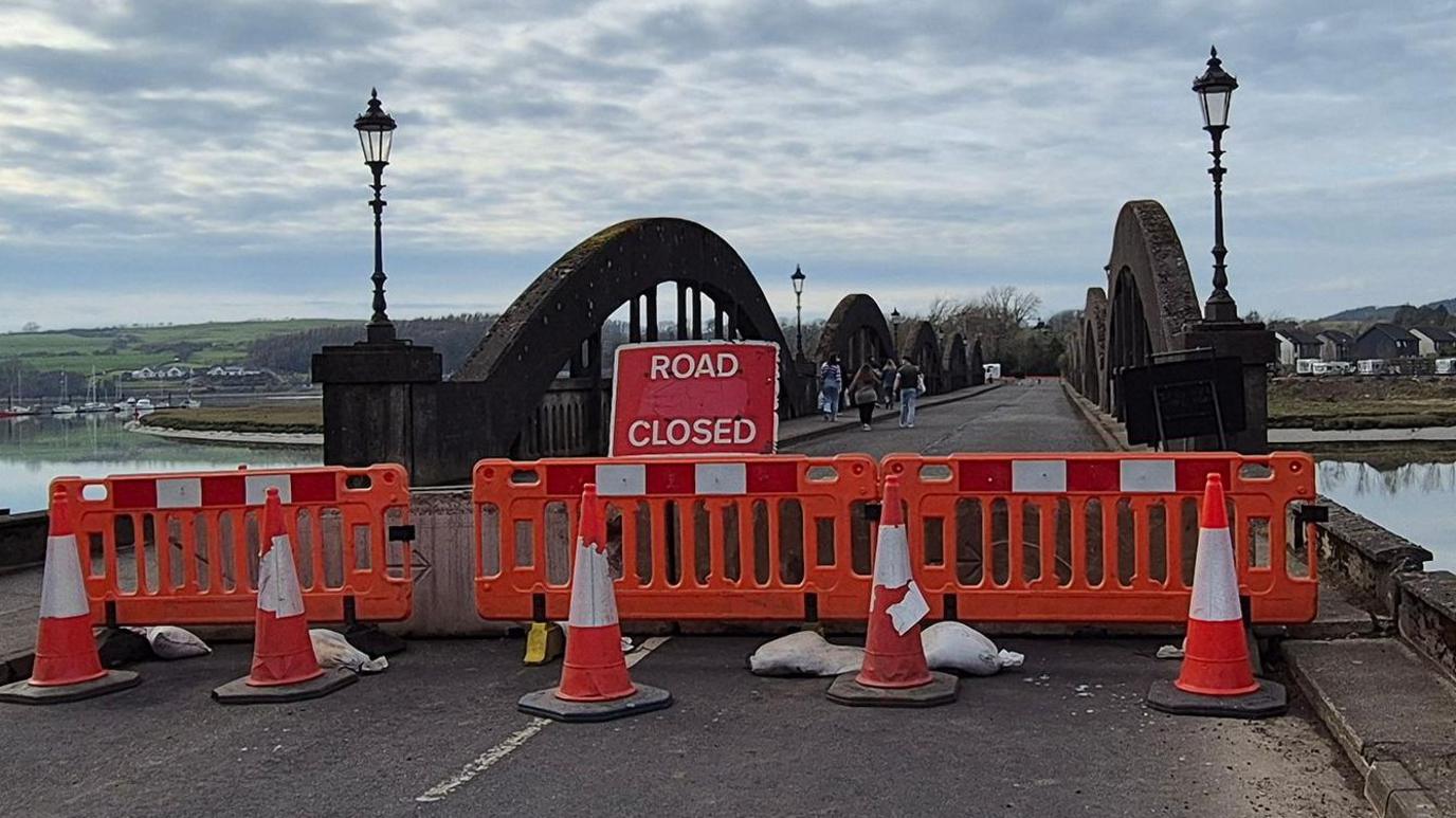 A road closed sign and orange barriers at one end of a bridge