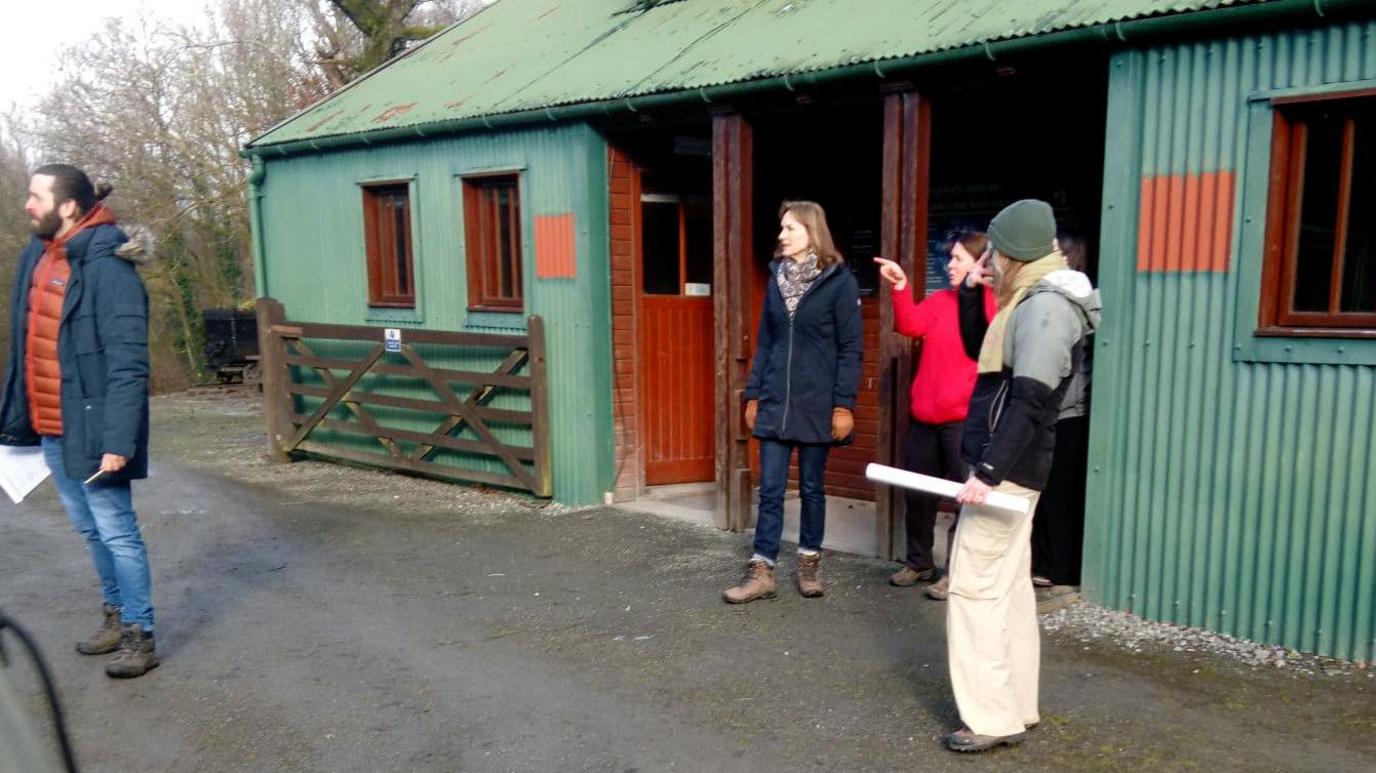 Fiona Bruce with members of the Fake or Fortune crew coming out of a green-painted metal barn. She is standing in a doorway on the right, wearing blue jeans, a blue jacket, gloves and her light brown hair is should-length. She is looking to the left, in the direction being pointed to by a woman in a red fleece. A member of the crew on the left wearing a hat, jacket and cream trousers is looking towards her. Another member of the crew with a dark beard and hair in a bun, wearing a blue jacket and jeans is on the far left. 