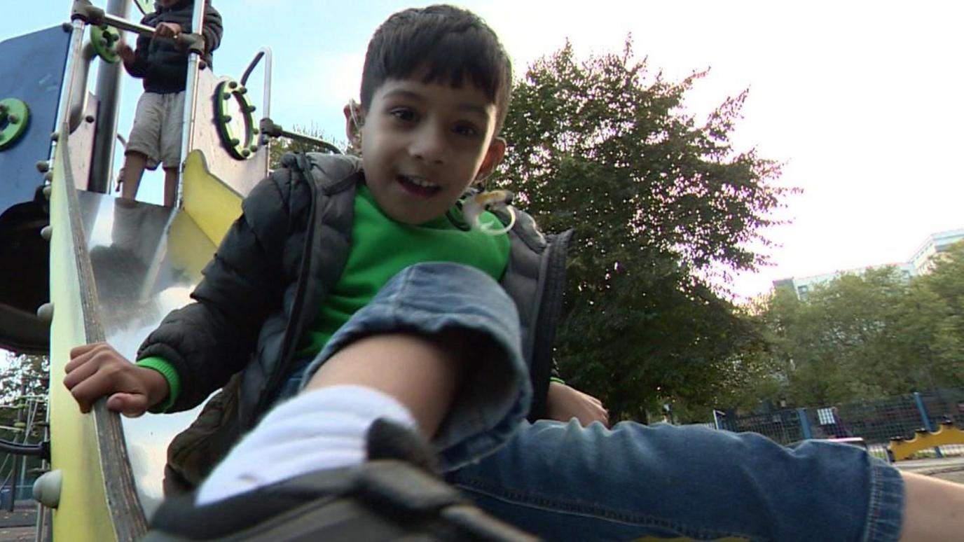 Muhaymin, a young boy wearing a black and grey coat and green jumper, and using a gold glittery hearing aid and cochlear implant, slides down a yellow slide in a children's play area