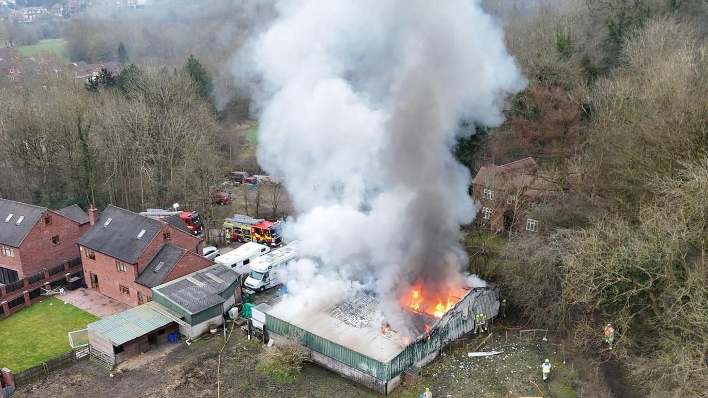 A drone shot of a single-storey brick-built factory unit on fire, surrounded by countryside and two homes 