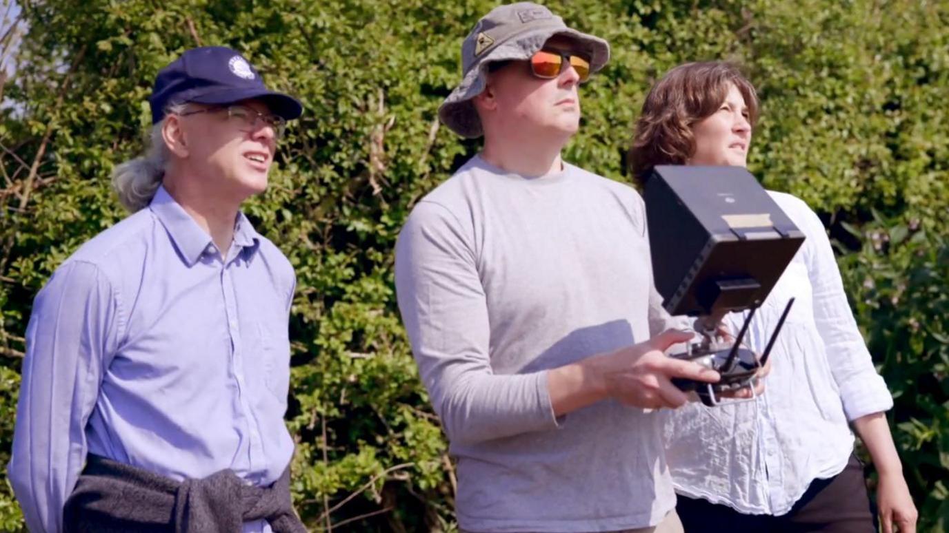 Three scientists are outdoors, looking up. One of them is holding a controller that has a camera attached to it. It's a sunny day.