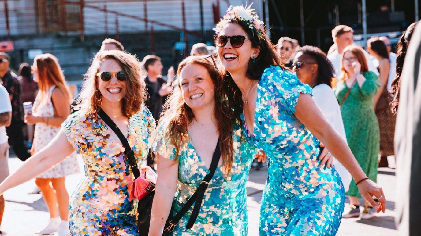 Three women in glittery jumpsuits smile at the camera as they wait for a concert to begin at Bristol Sounds 2024