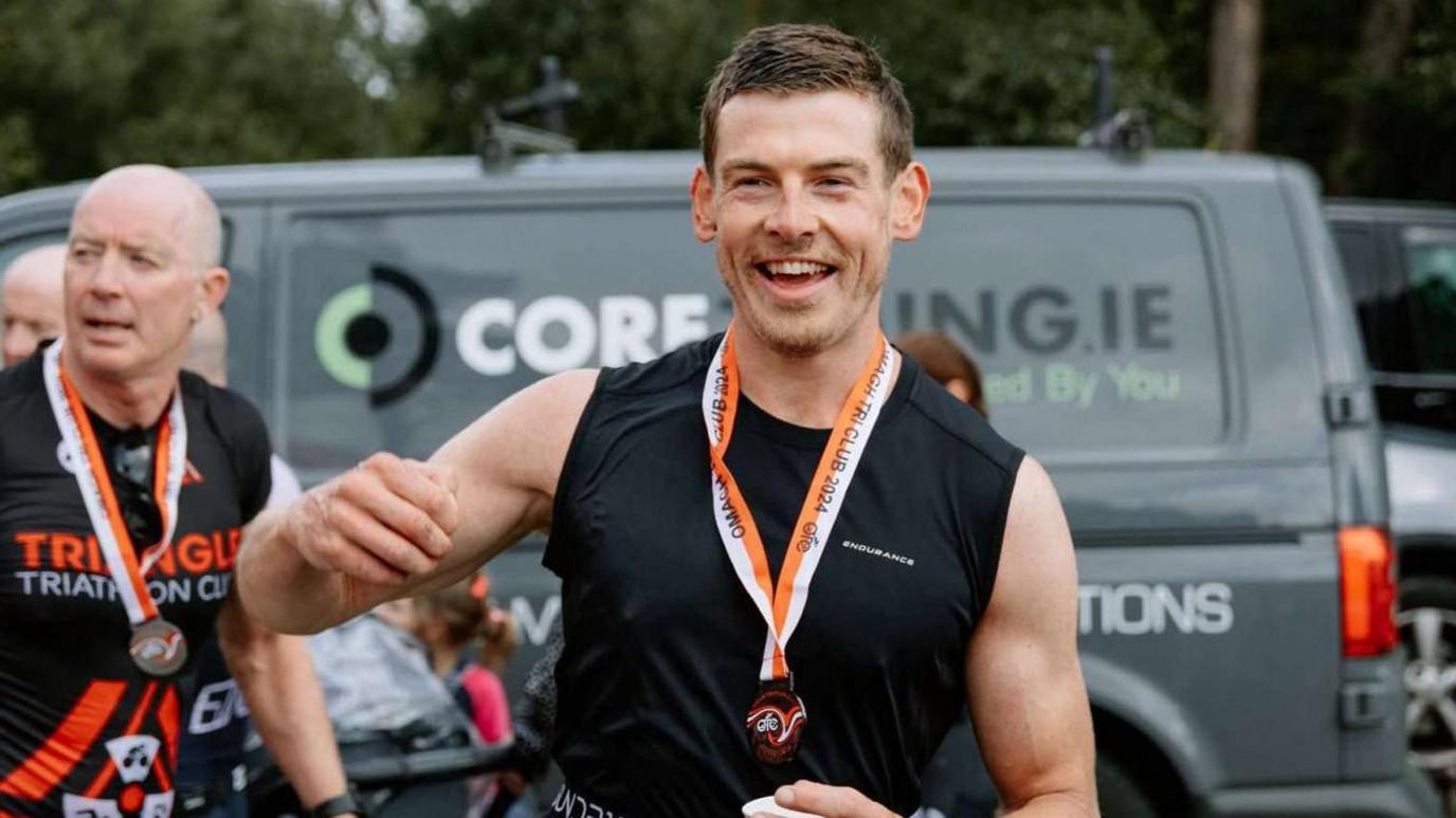 A young man with brown hair is running and smiling off to the side of the camera. He is wearing a black running vest and has an orange and white medal around his neck. A silver van is in the background. Another man who has a shaved head is to the left of him. He is wearing the same medal.