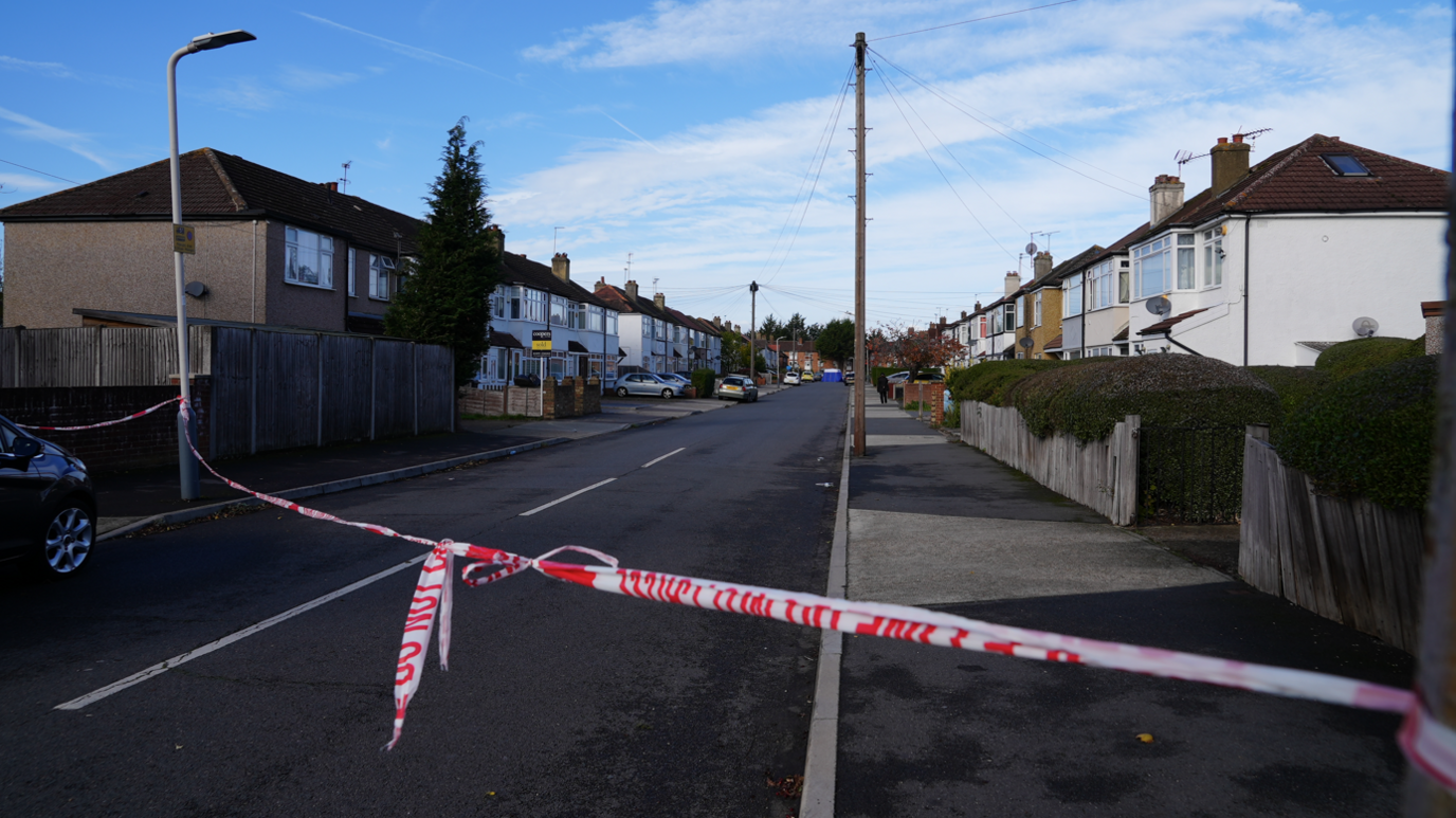 Red and white police tape is tied on lampposts across a road which has houses running alongside either side of it