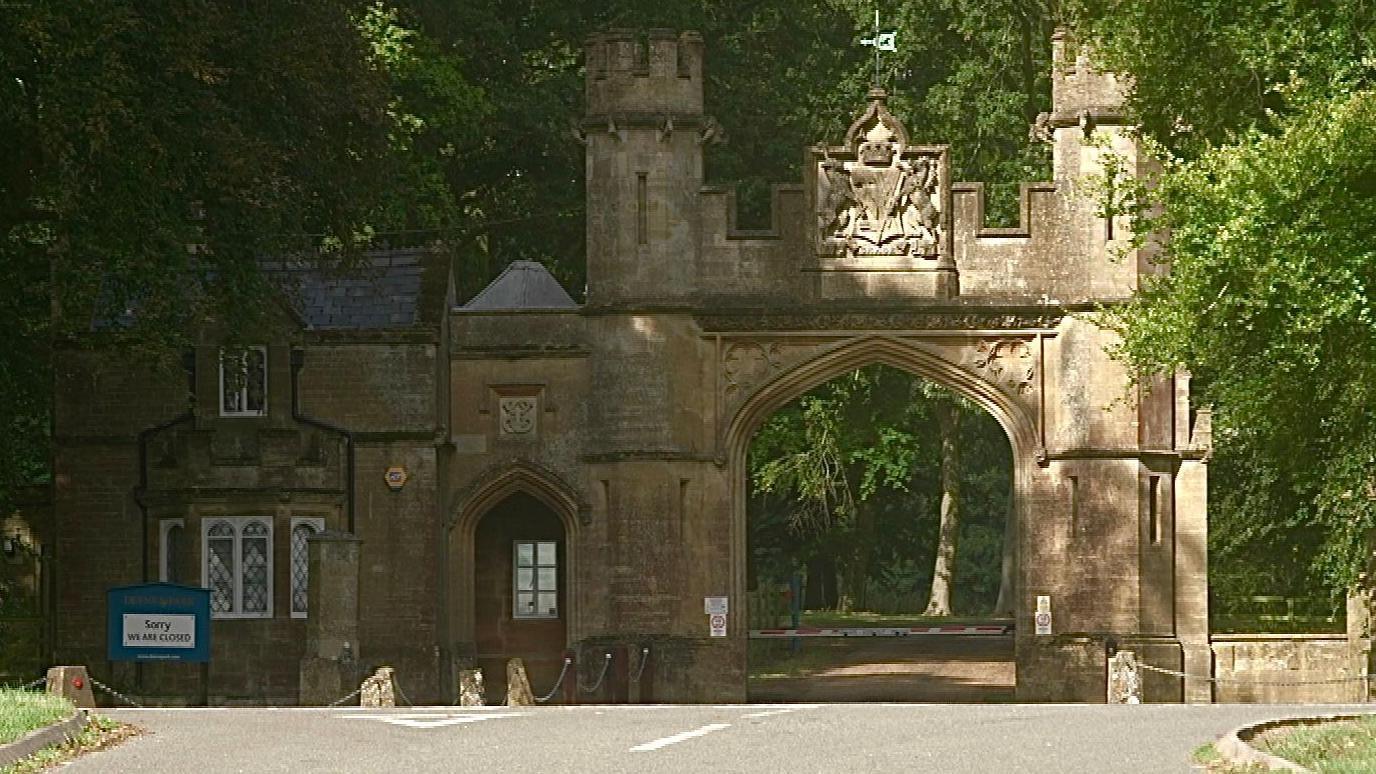 The gate house of Deene Park, a stately home, built from stone. The gate half, on the right with the road entry, has two mini castellated towers on either side. The house side on the left is a small cottage with diamond-paned windows. A road is in front and trees surround it