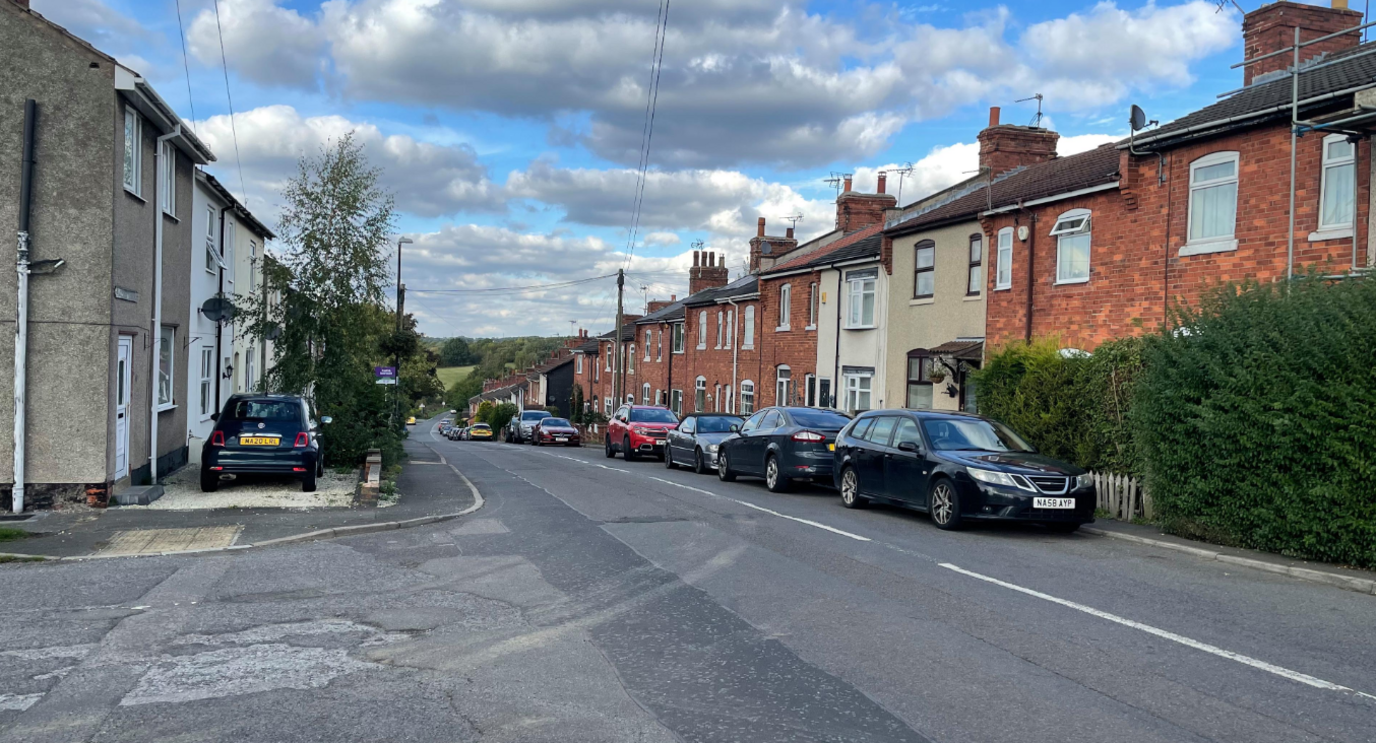 A view of Midland Terrace in Westhouses, Derbyshire. Several cars can be seen parked either side of the road with terraced houses on both sides 