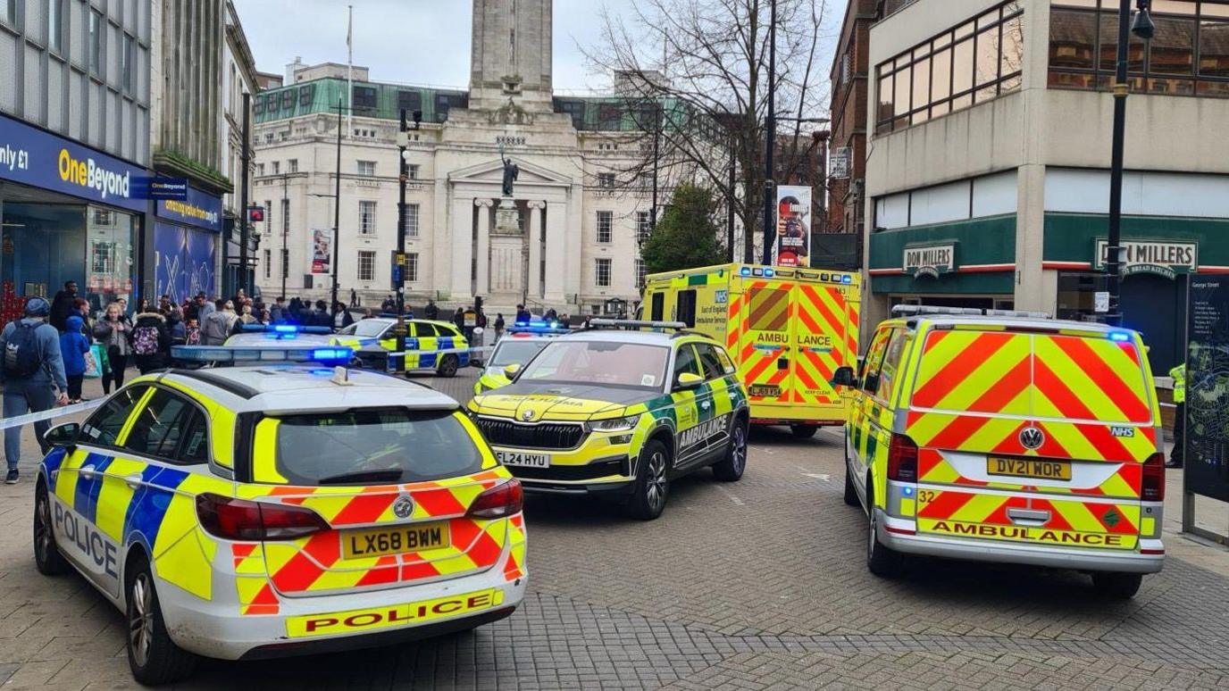 Police cars and ambulances parked on a wide pedestrianised street in the centre of Luton. They are inside a cordon. There is blue-and-white police tape sealing that part of the pavement off. There are shoppers in the street and Luton town hall can be seen behind the emergency vehicles.