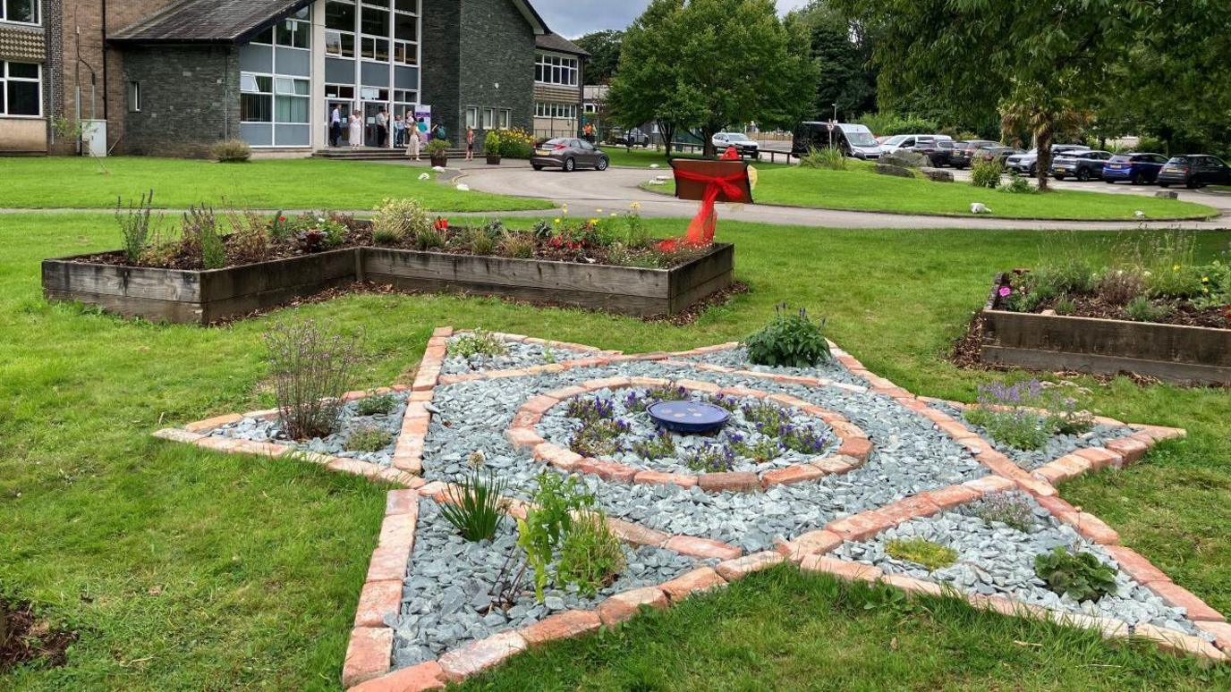 A memorial garden. Flower in wooden planters mark out a square in the grass. In the centre, slate and brick is arranged to a six-pointed Star of David. A plaque is wrapped in red ribbon.